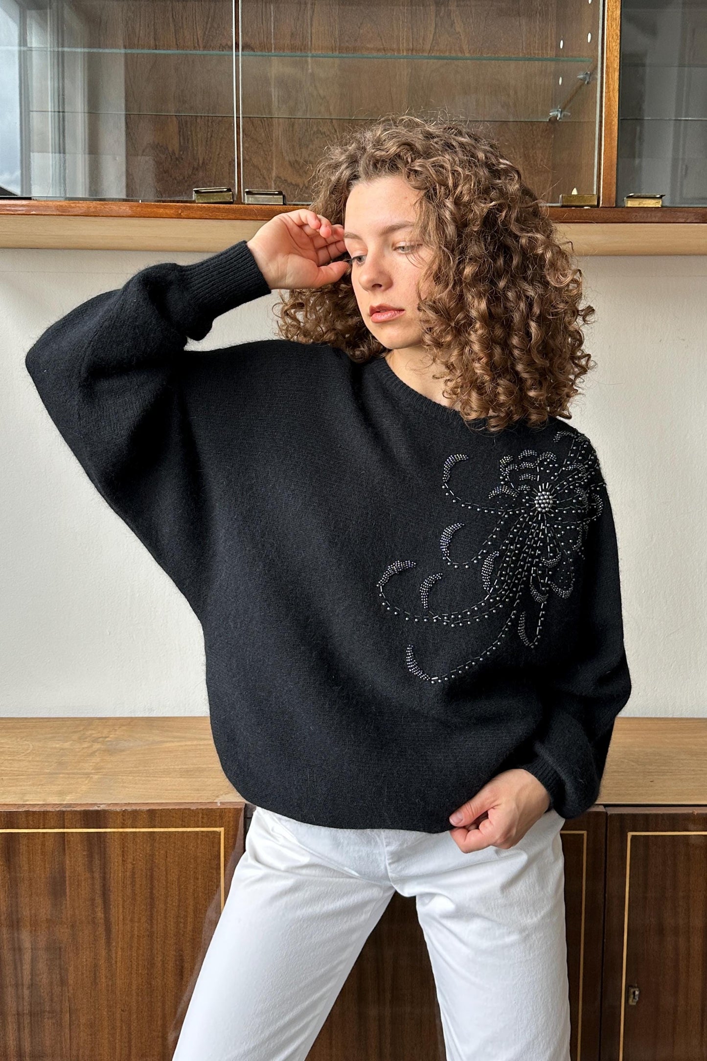 A woman with curly hair wearing a black sweater with a floral design stands in front of a glass display case.