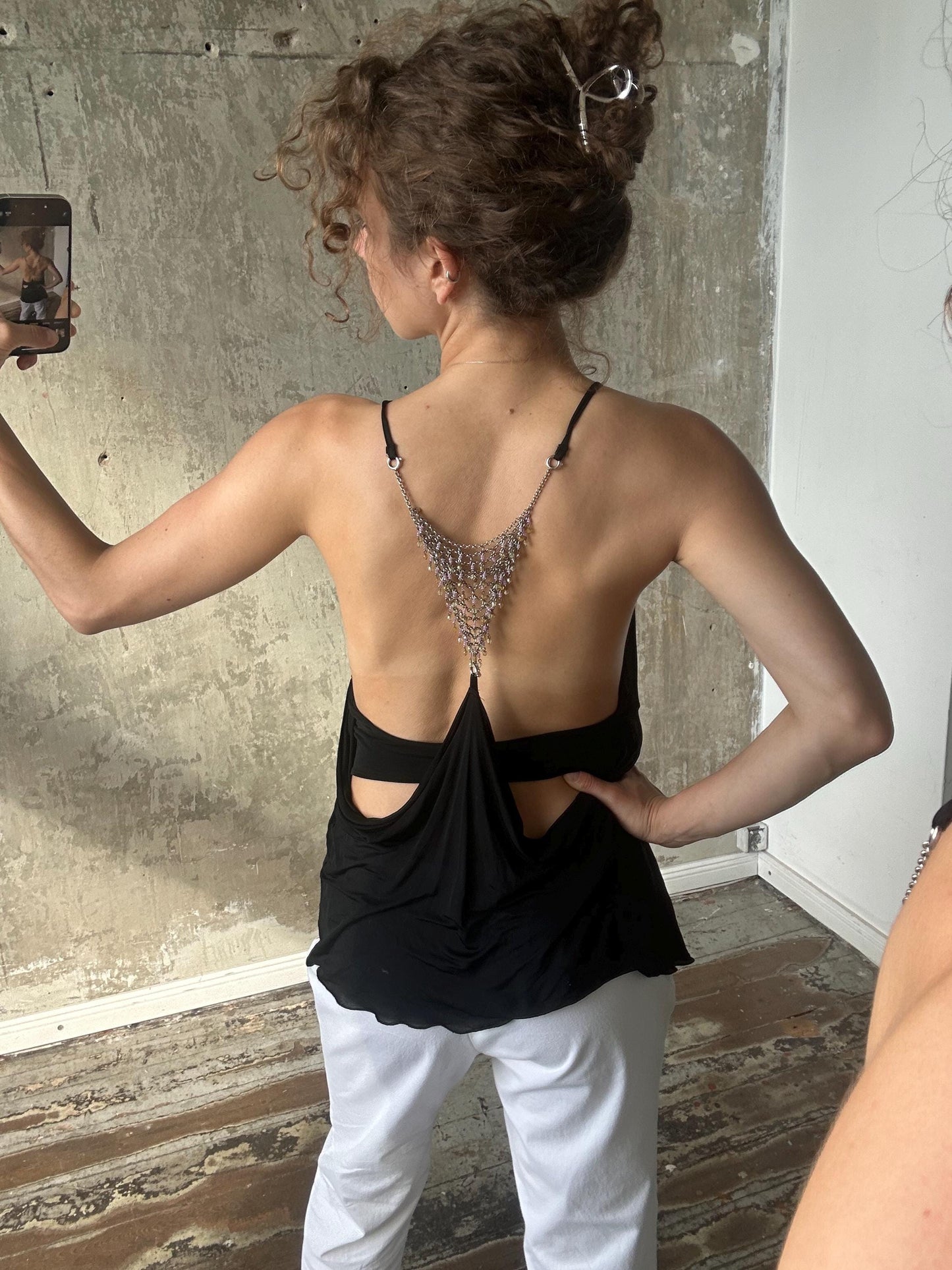 a woman taking a selfie with a cell phone, wearing a black top with a backless design and white pants.