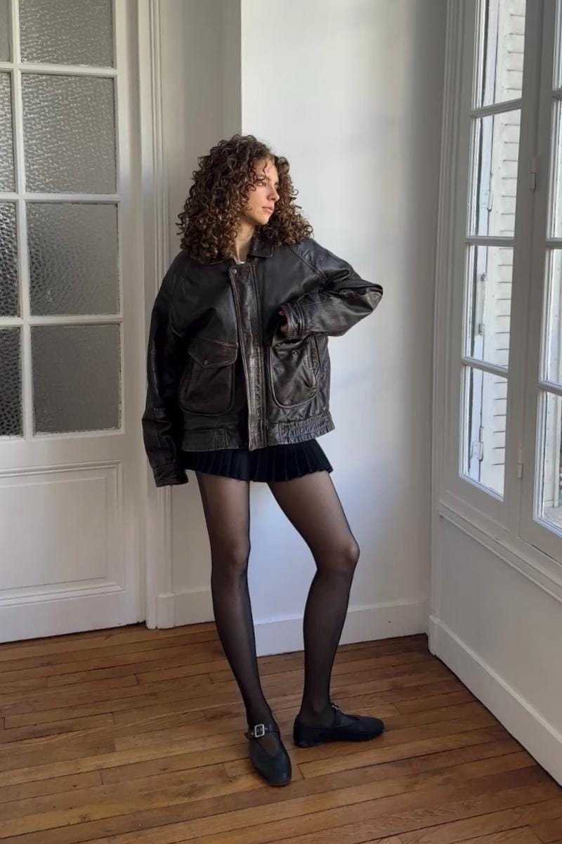 A woman with curly hair stands in a room, wearing a black leather jacket and a short skirt.