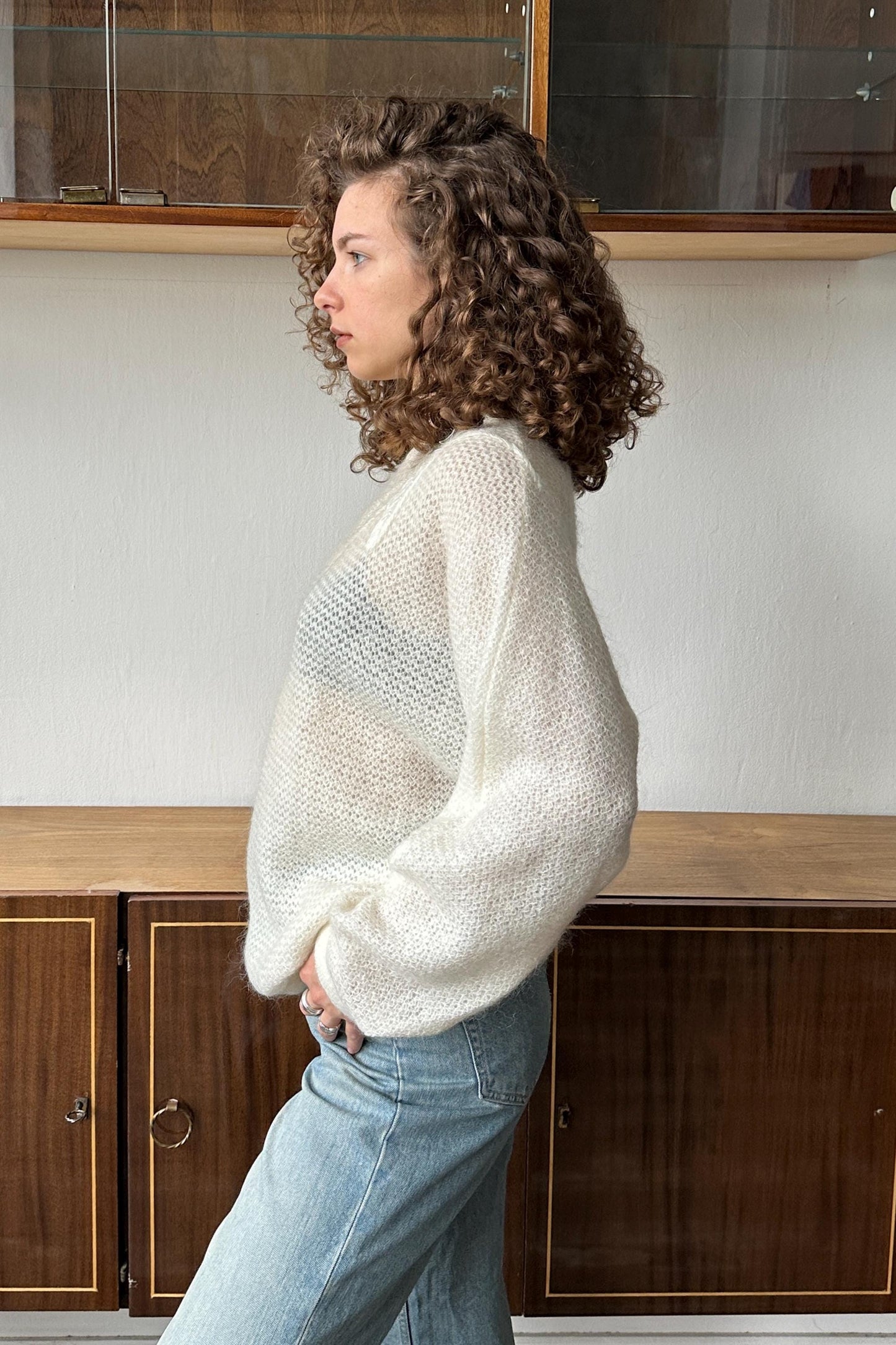 A woman with curly hair wearing a white sweater and blue jeans stands in a room with wooden cabinets in the background.