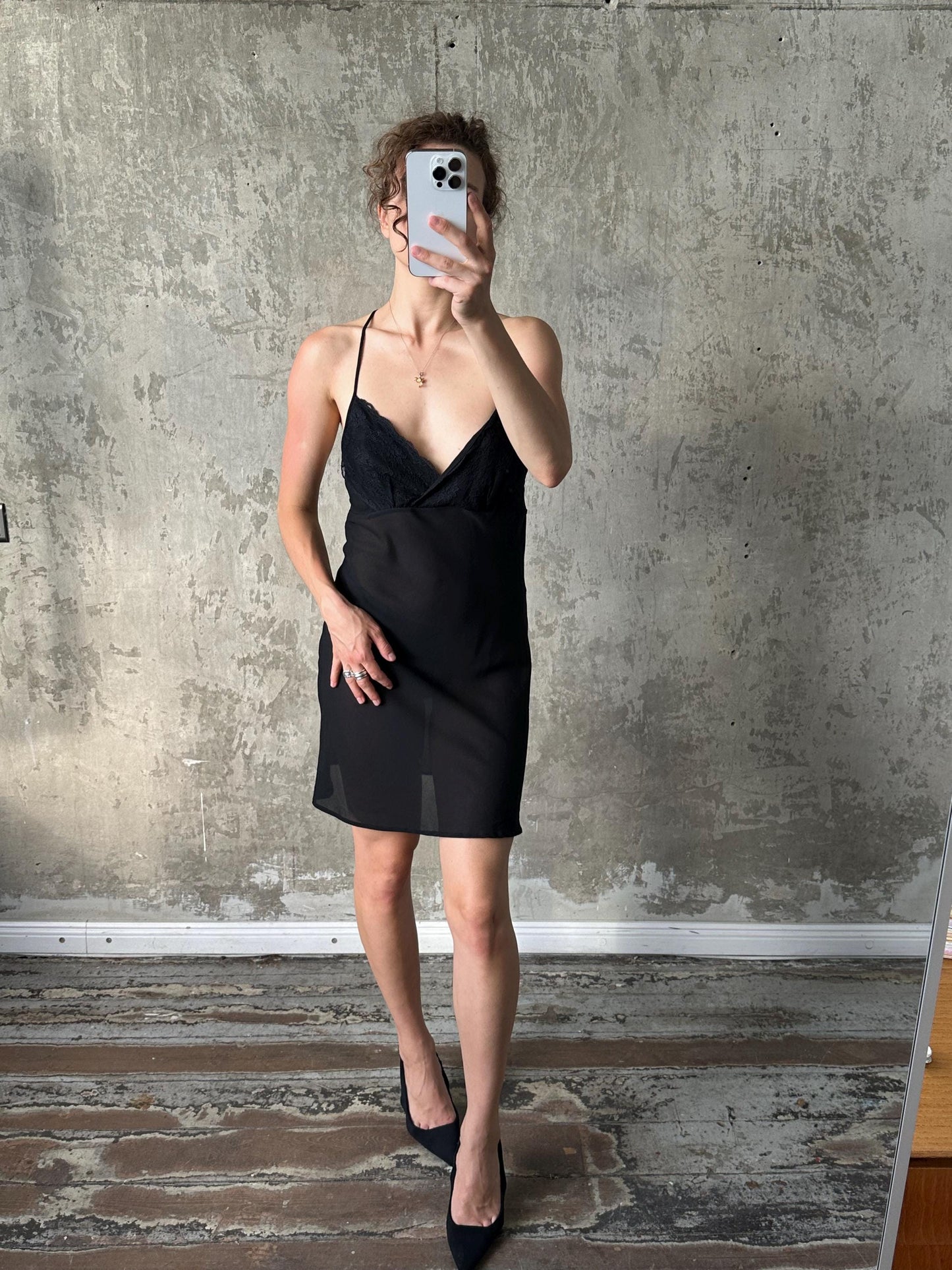 A woman in a black dress is taking a selfie with a smartphone, standing in front of a concrete wall.