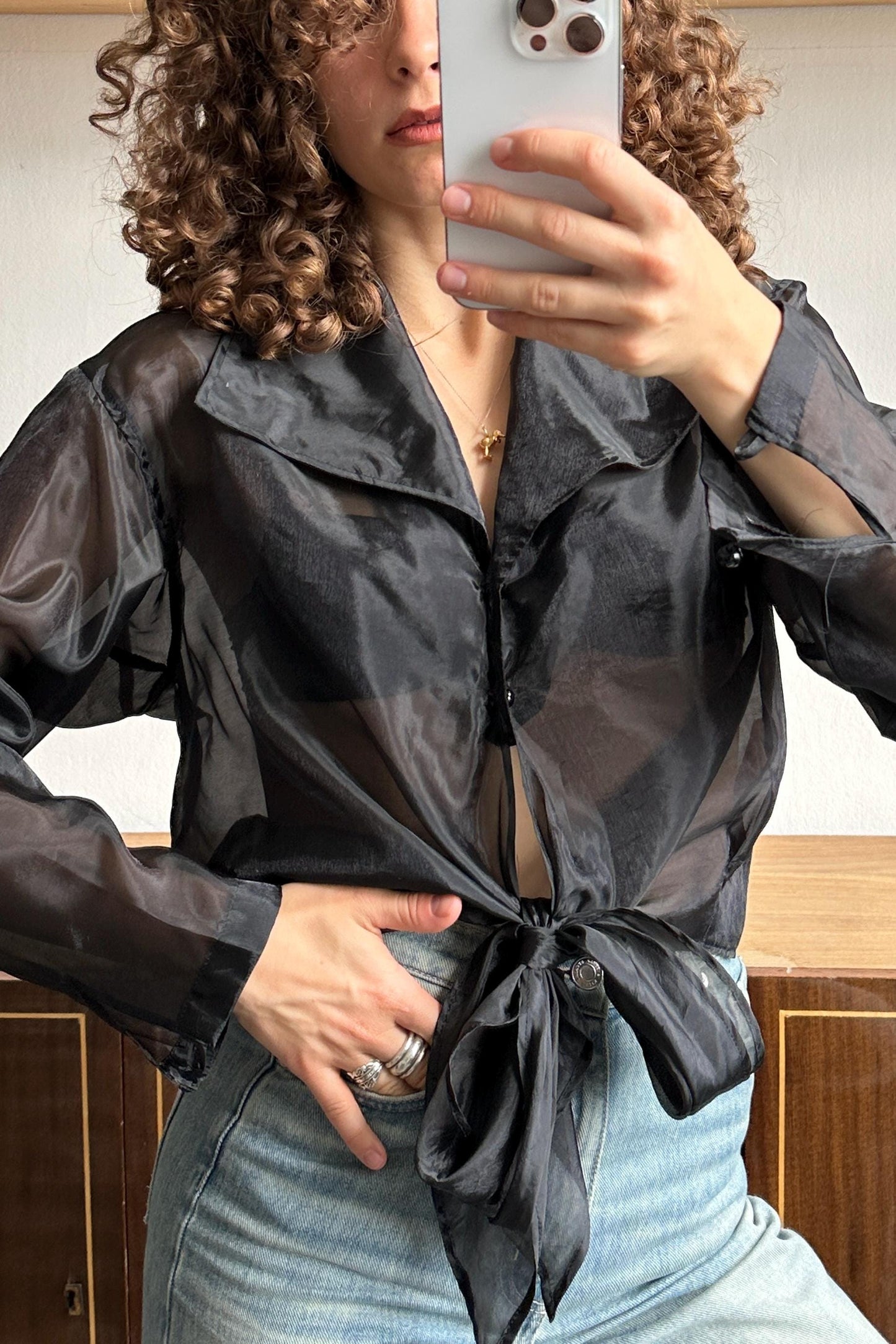 A woman with curly hair is taking a selfie with a smartphone, wearing a black blouse with a tie and blue jeans.