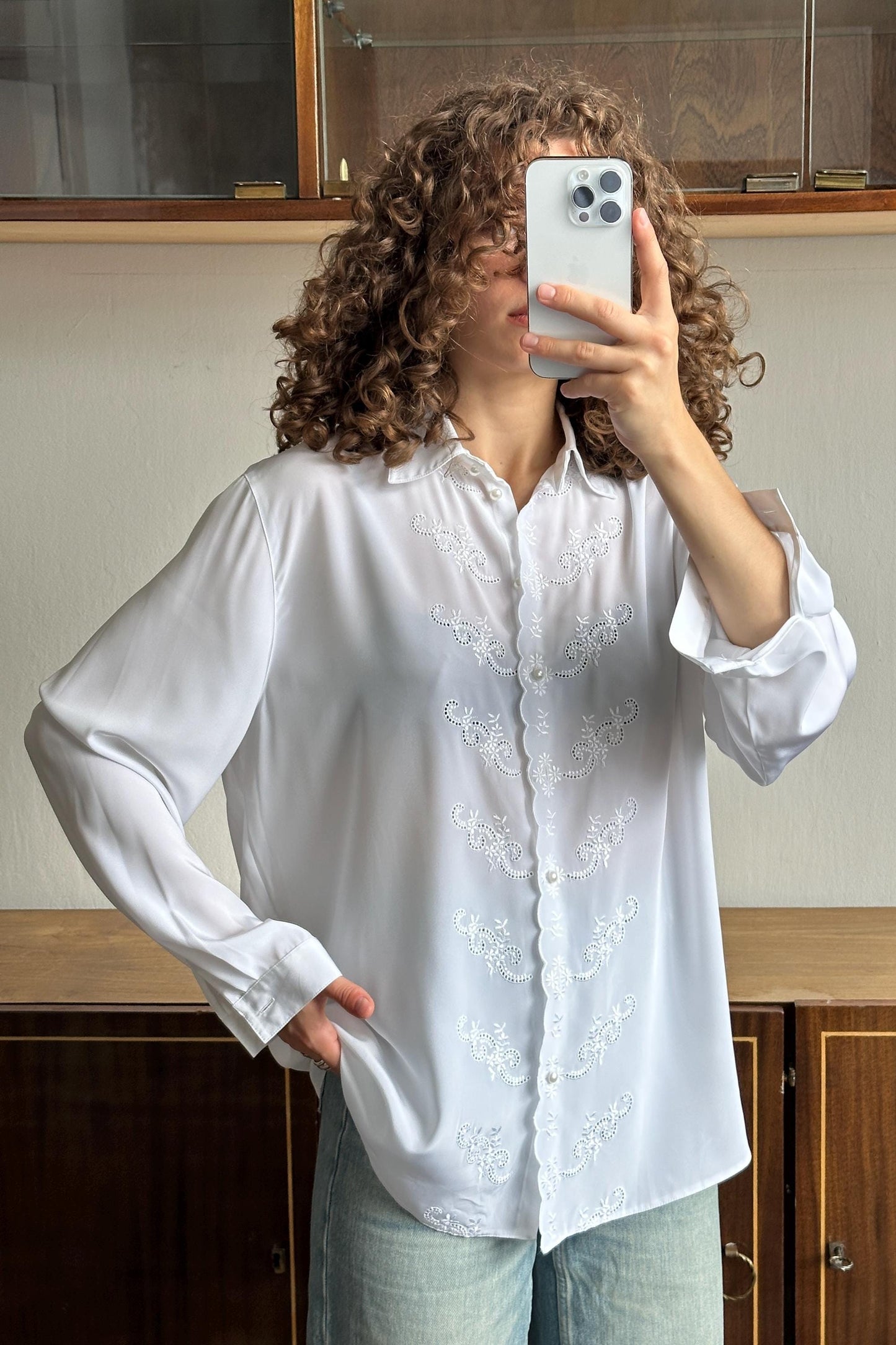 A woman with curly hair is taking a selfie with a white smartphone, wearing a white blouse with embroidered designs.