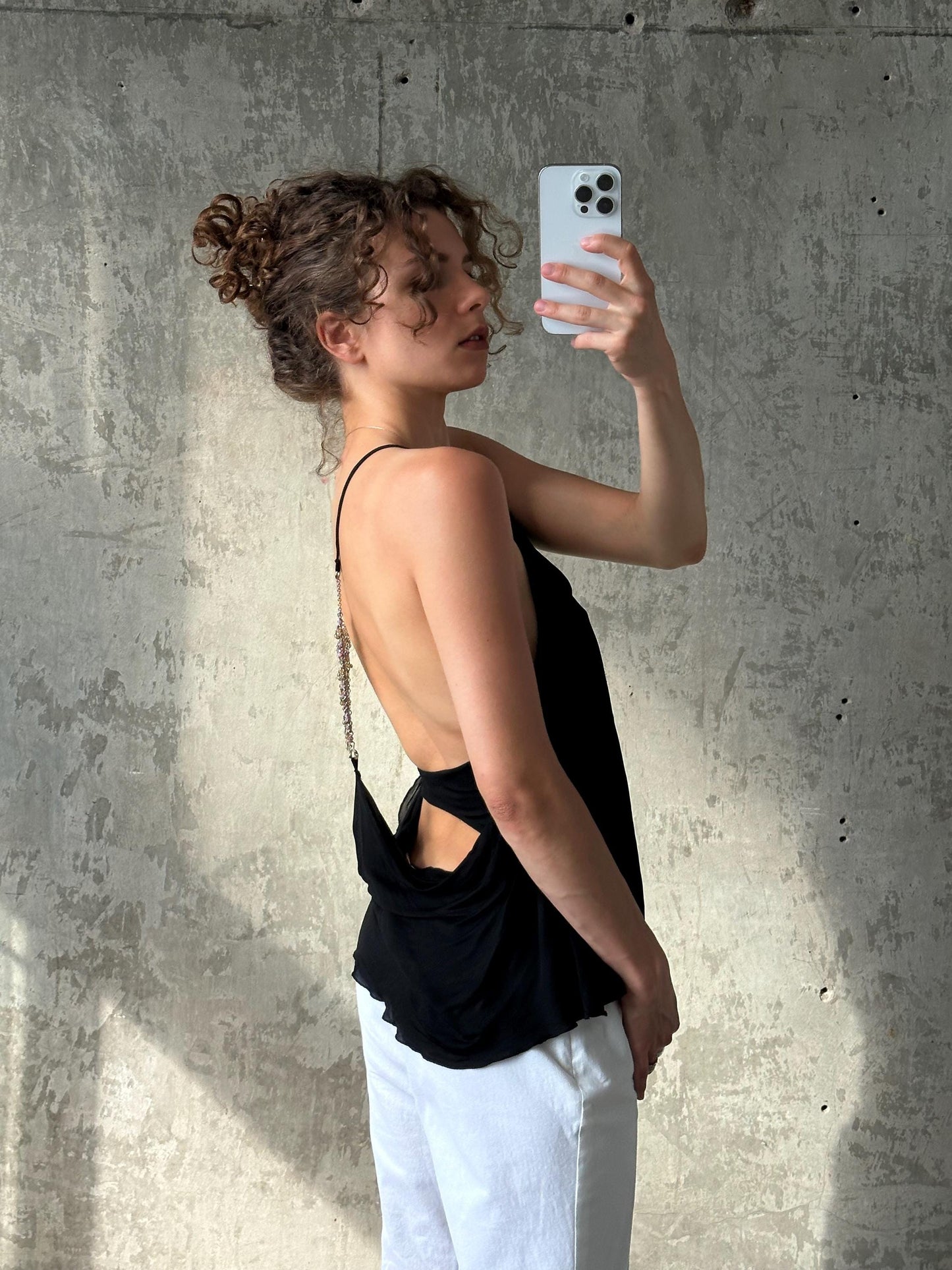 A woman with curly hair is taking a selfie with a white phone, standing in front of a concrete wall.