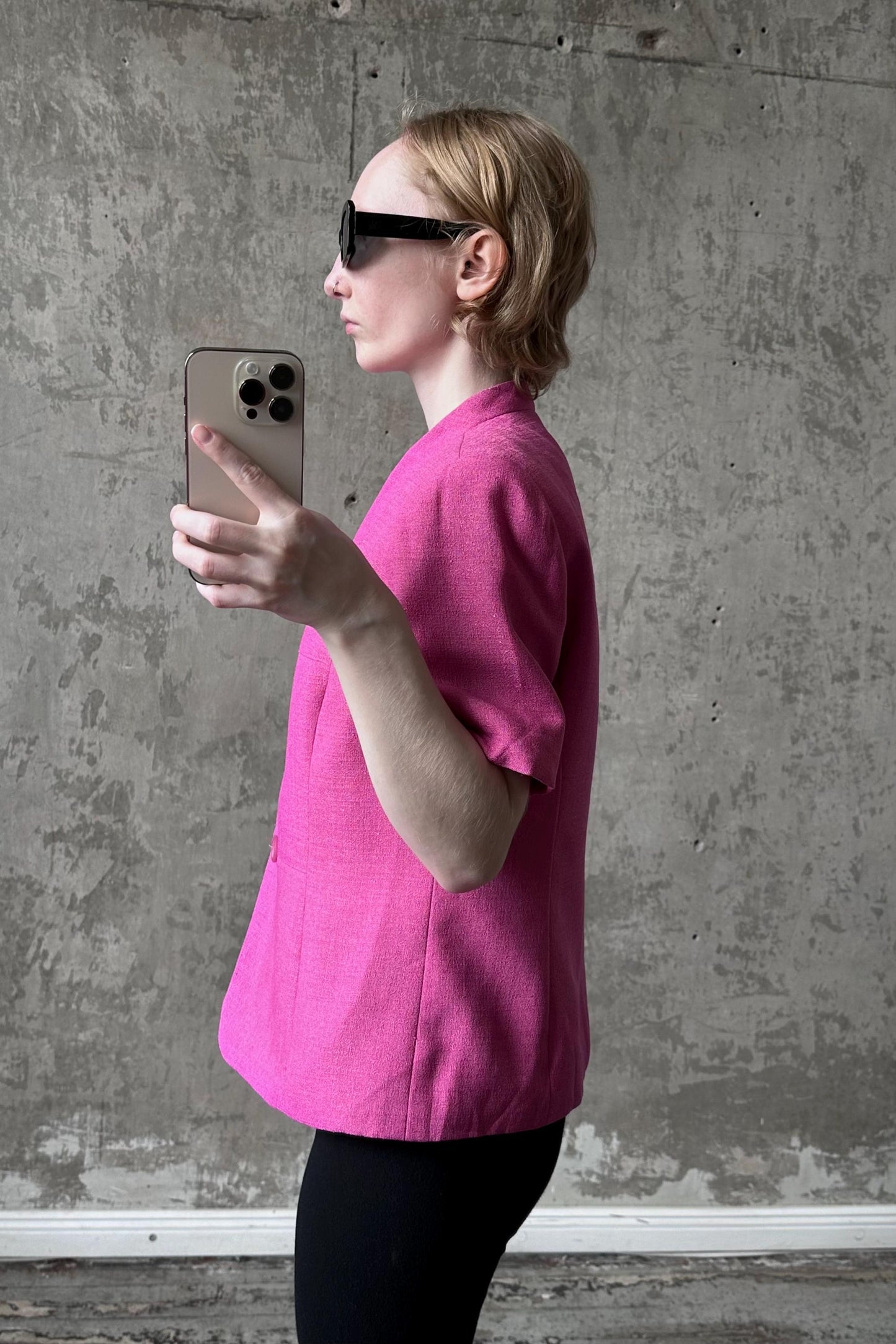 A woman in a pink blazer is holding a smartphone and taking a selfie in front of a concrete wall.