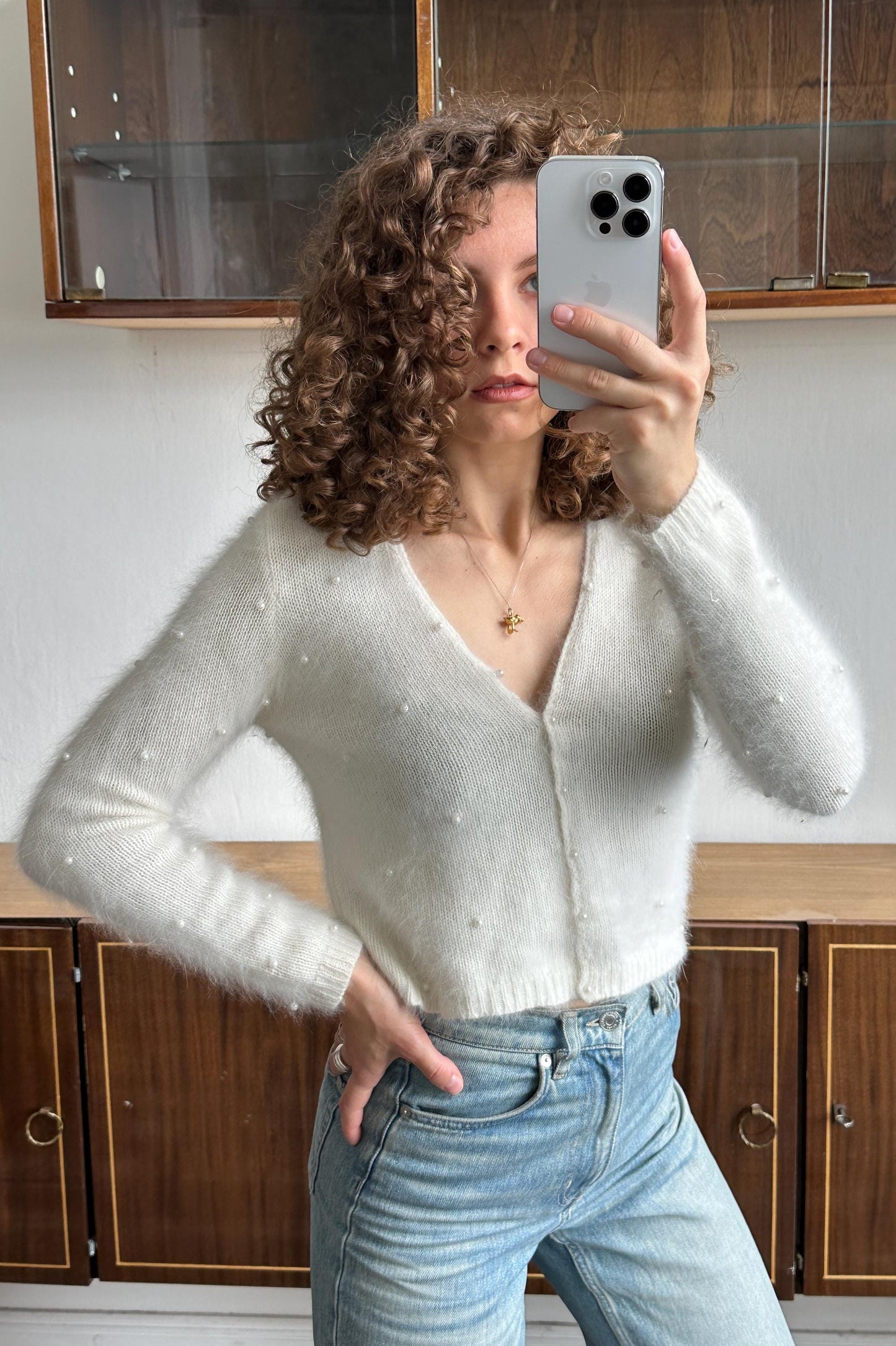 A woman with curly hair is taking a selfie with her iPhone, holding the device up to her face. She is wearing a white sweater with polka dots and blue jeans.