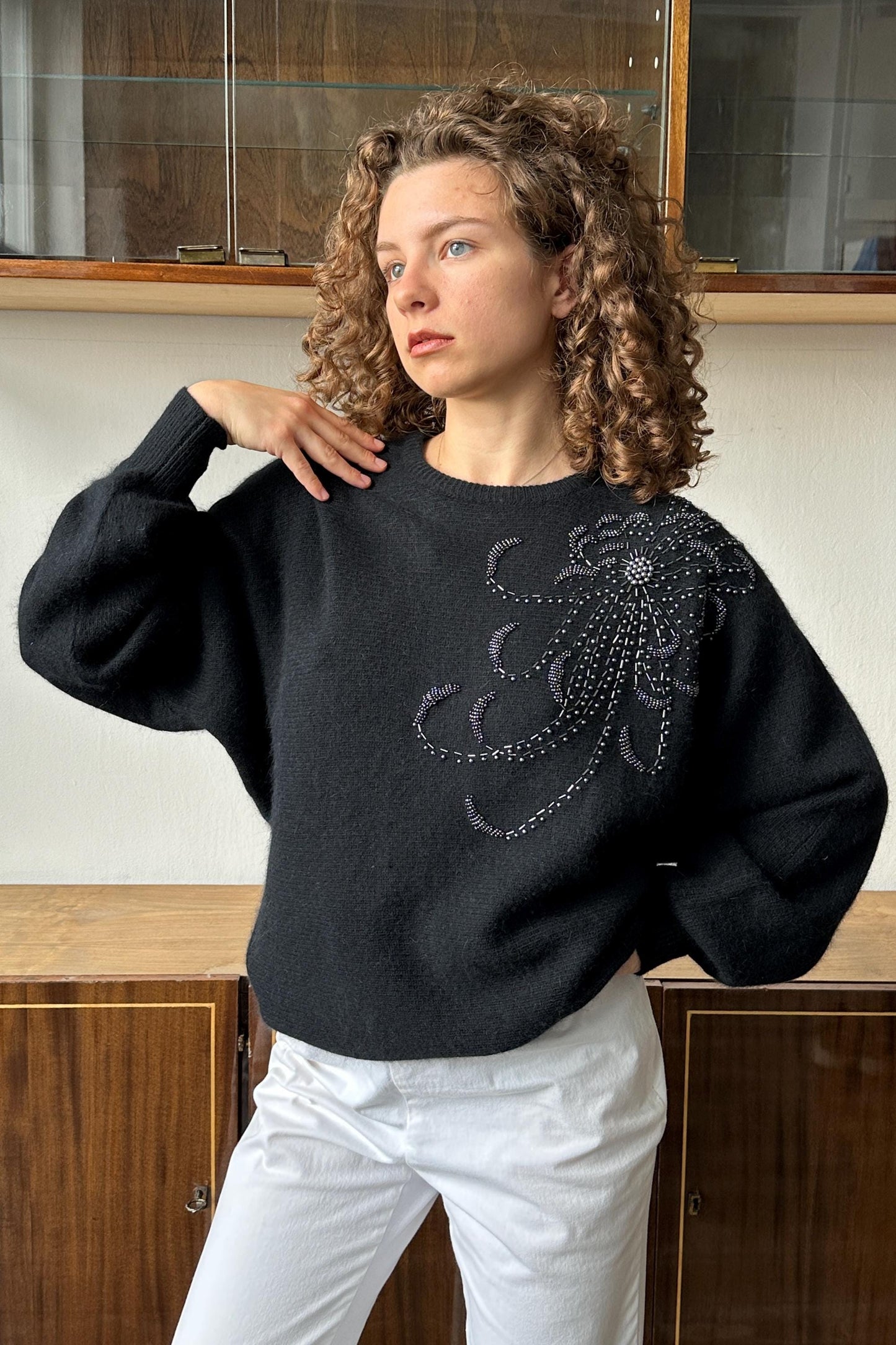 A woman with curly hair wearing a black sweater with a floral embroidered design stands in front of a wooden cabinet.