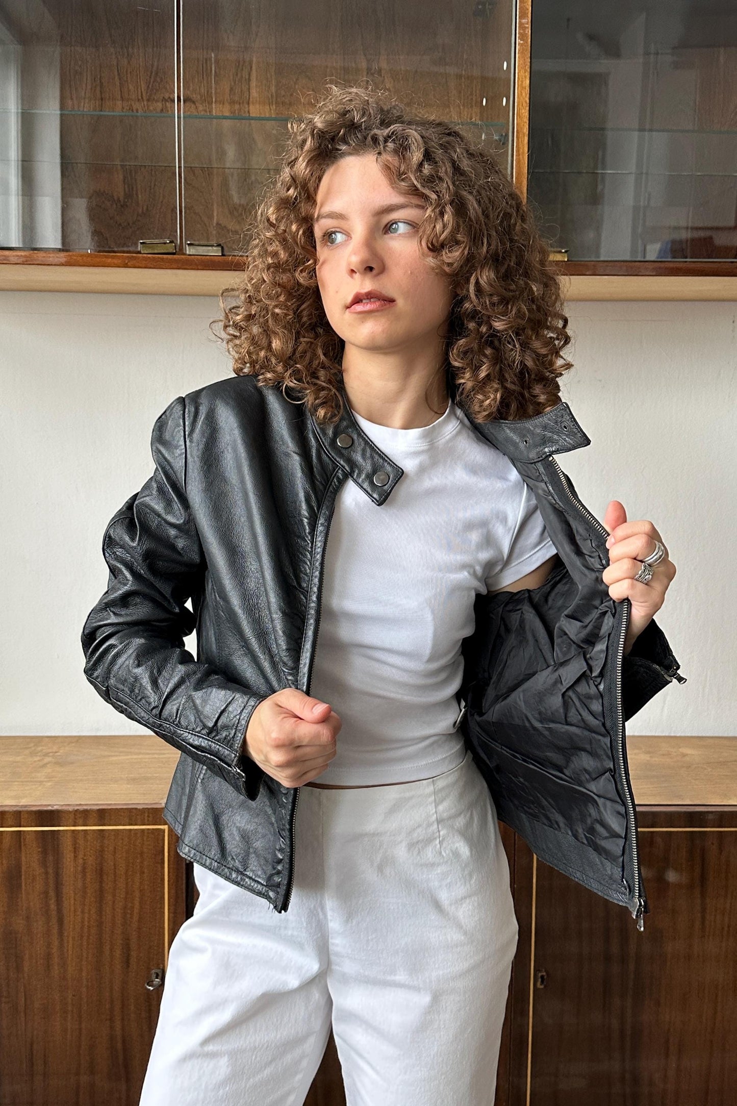 A young woman with curly hair wearing a black leather jacket and white pants stands in front of a wooden cabinet.