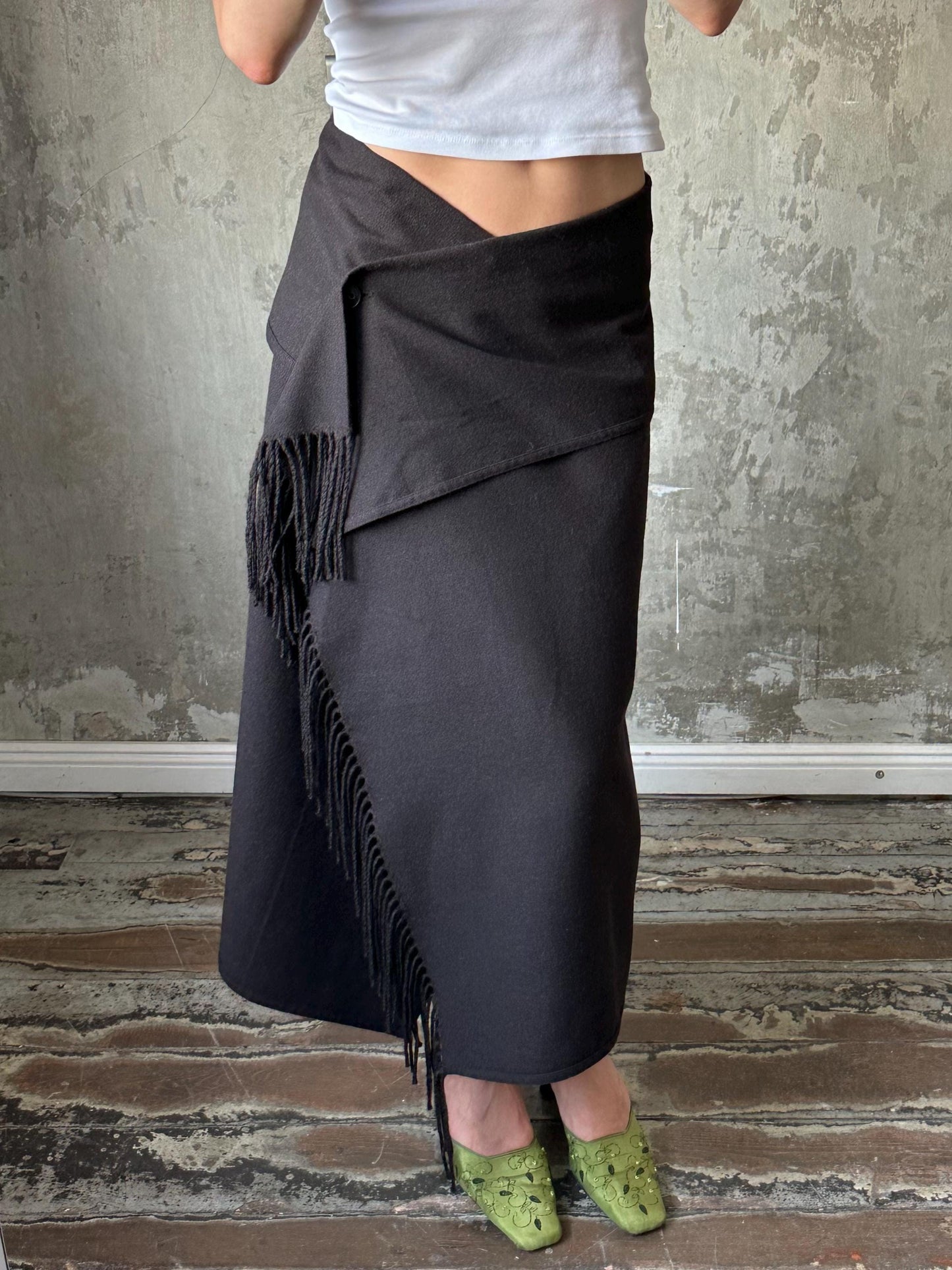 A person wearing a black skirt with fringe details and green shoes stands in front of a concrete wall.