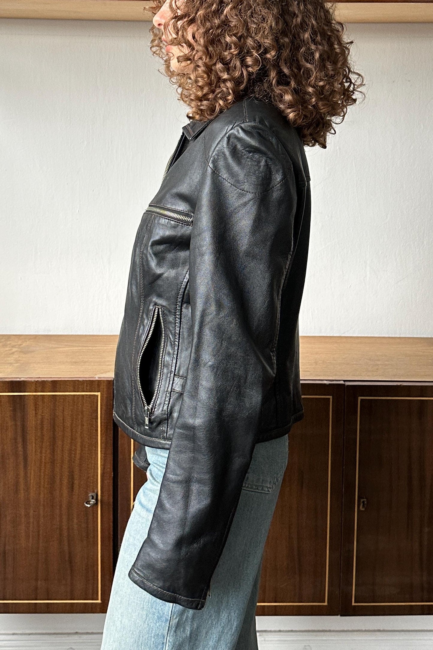 A woman with curly hair wearing a black leather jacket stands in front of a wooden cabinet.
