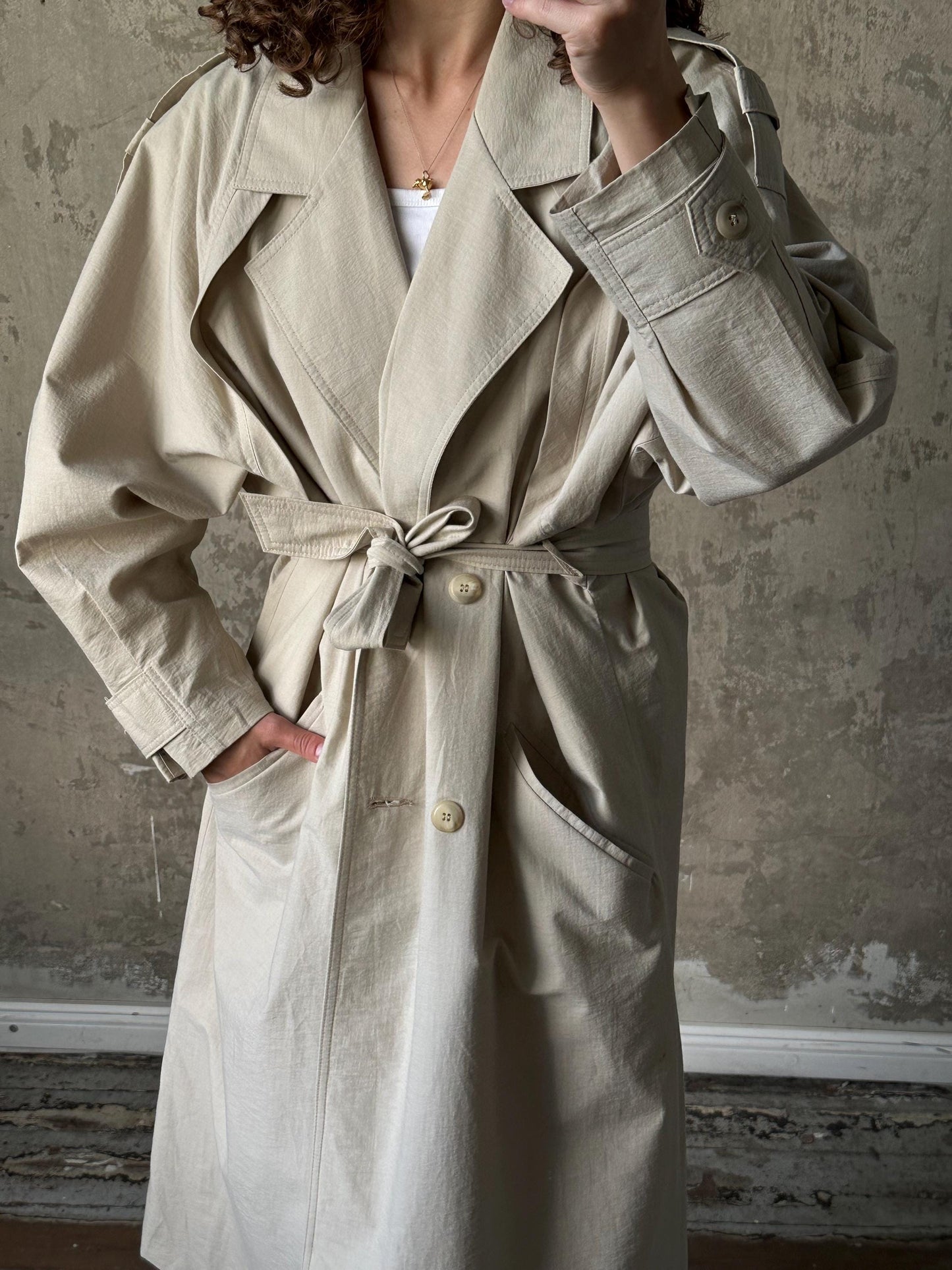 A person wearing a beige trench coat with a tie, standing against a concrete wall.