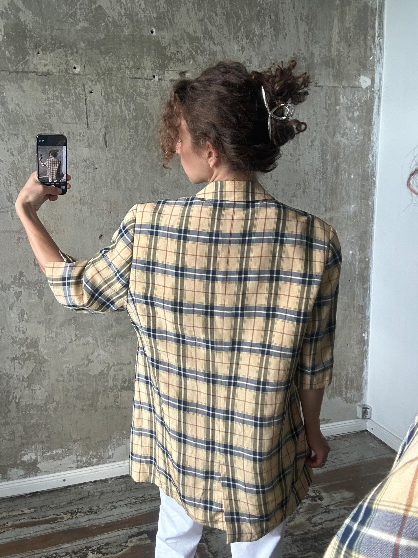 A person is taking a selfie with a smartphone, wearing a plaid shirt and having their hair in a bun.