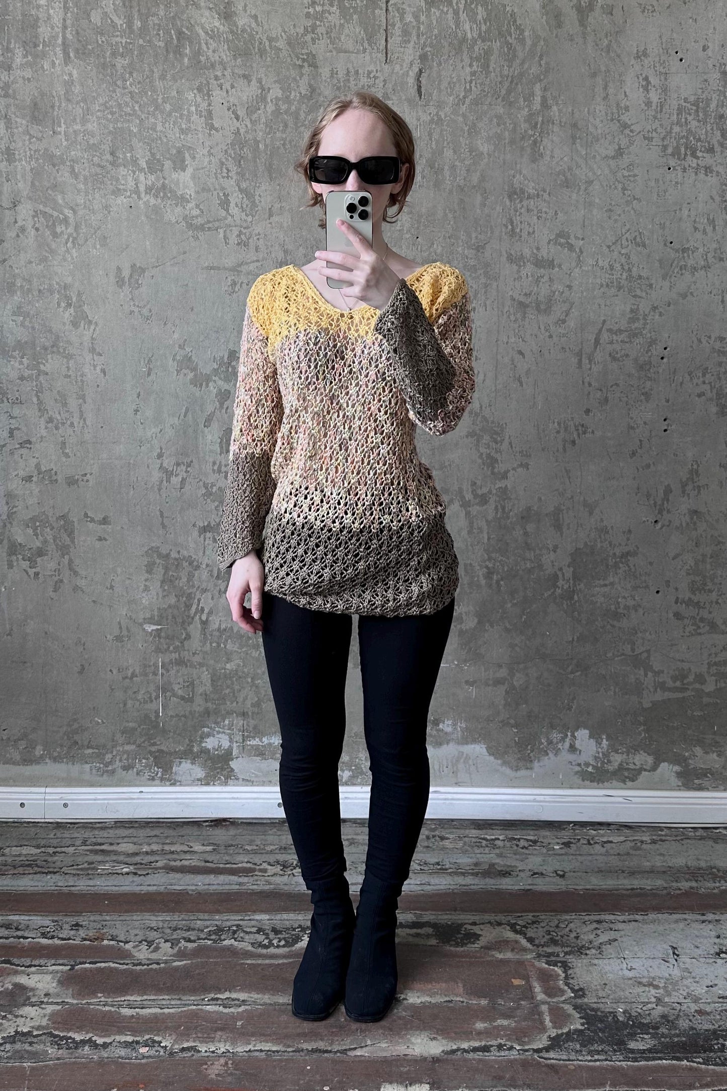 A woman wearing a yellow and brown patterned top, black leggings, and black boots is taking a selfie with a smartphone against a concrete wall.