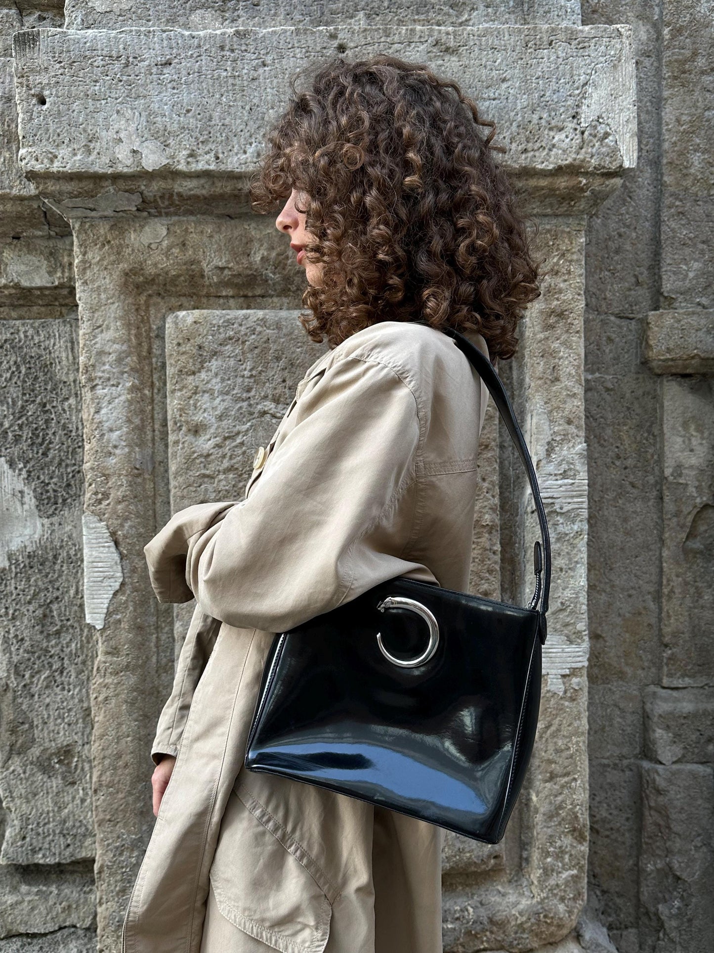 A woman with curly hair stands in front of a stone wall, wearing a beige coat and carrying a black purse with a silver circular logo.