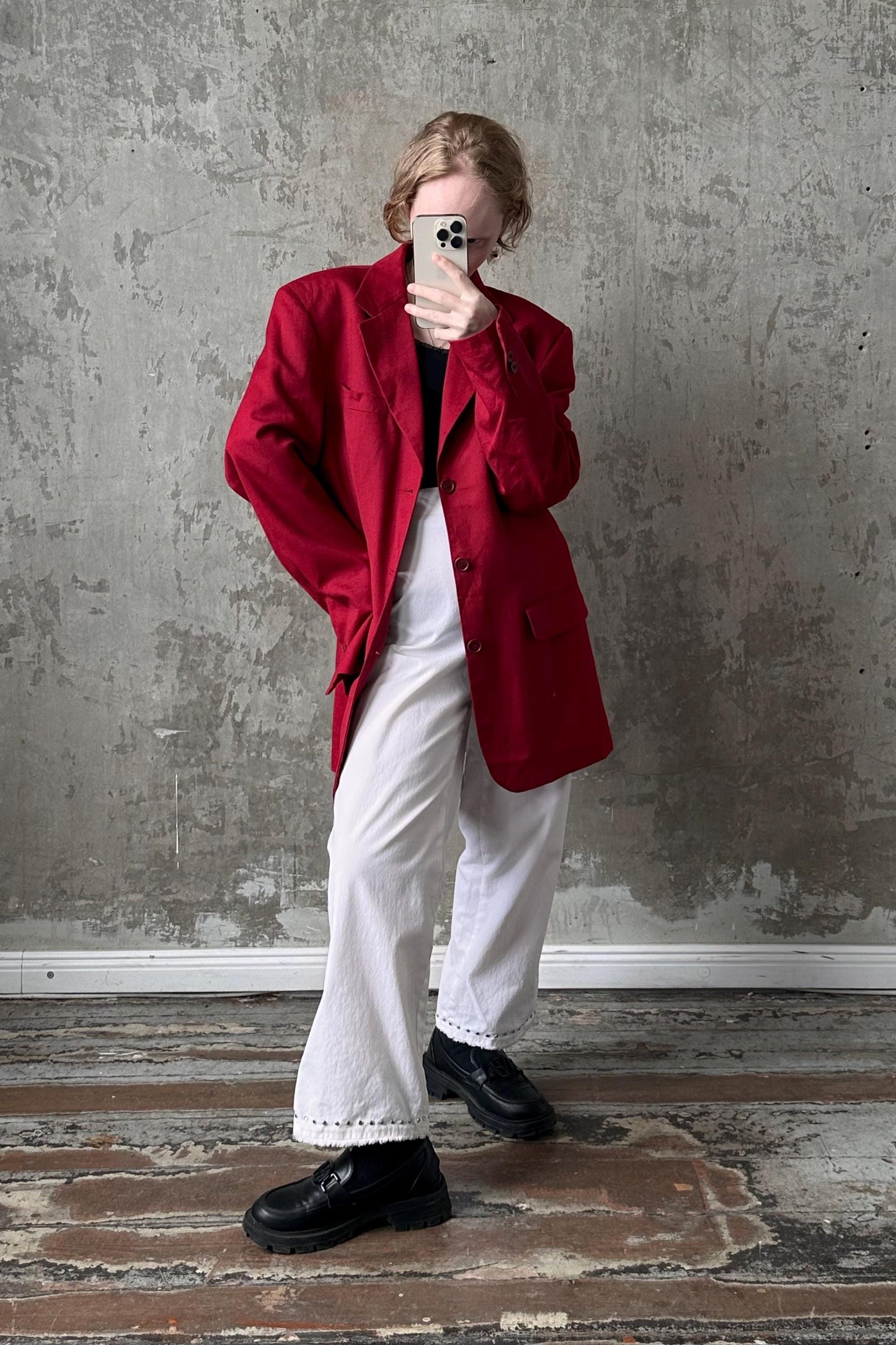 A person wearing a red jacket, white pants, and black shoes is taking a selfie with a smartphone against a concrete wall.