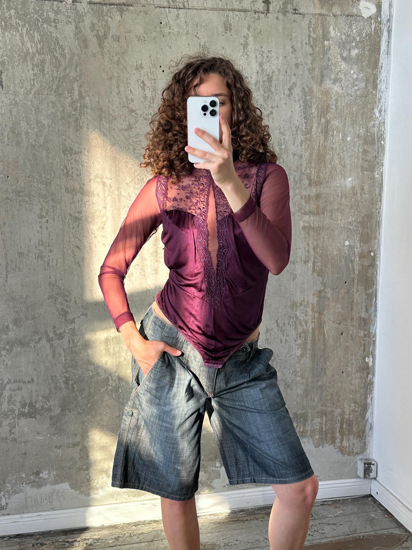 A woman with curly hair is taking a selfie with a white smartphone, wearing a purple top and blue jeans.