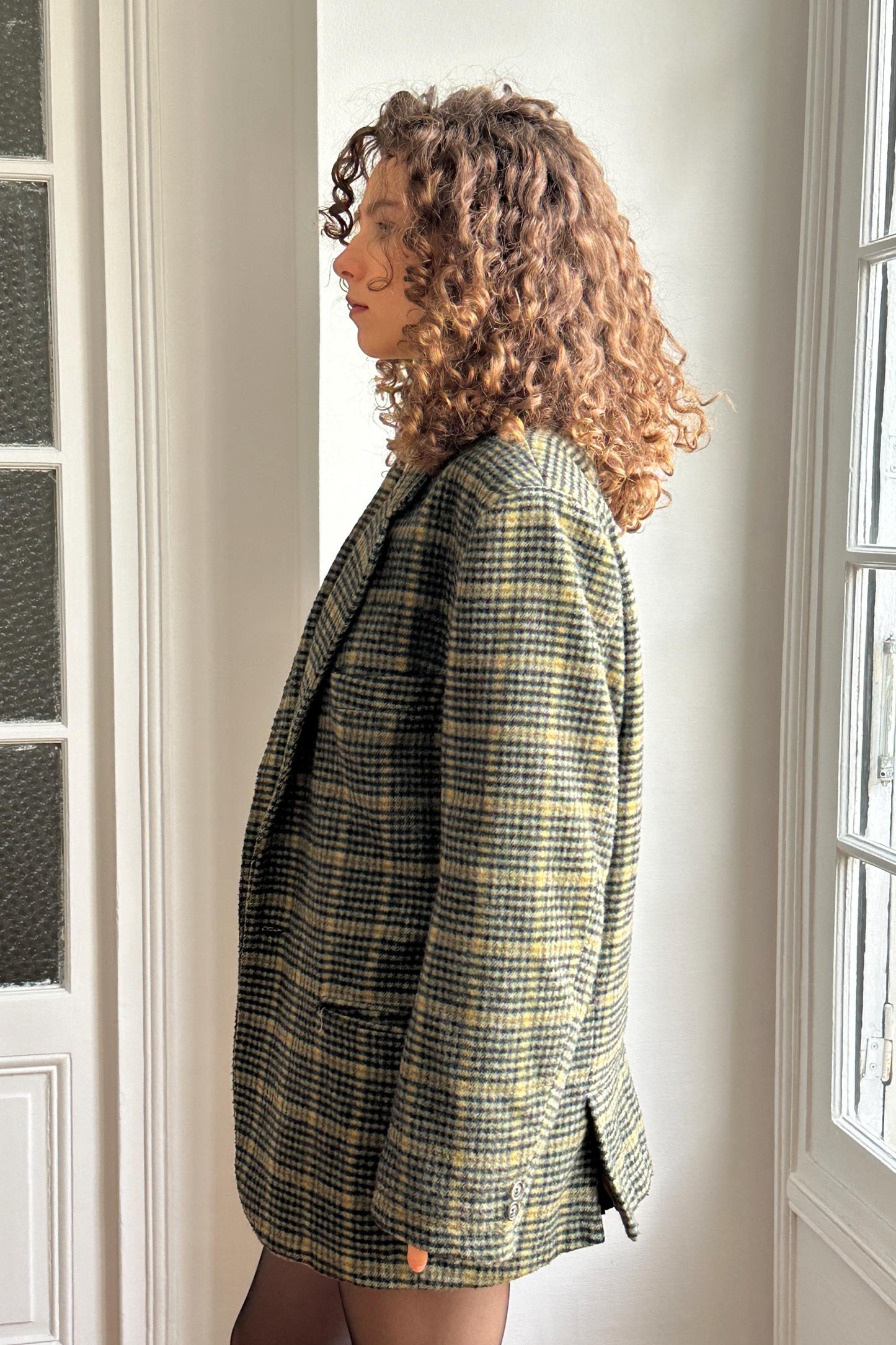 A woman with curly hair wearing a plaid jacket stands in a room with a window.