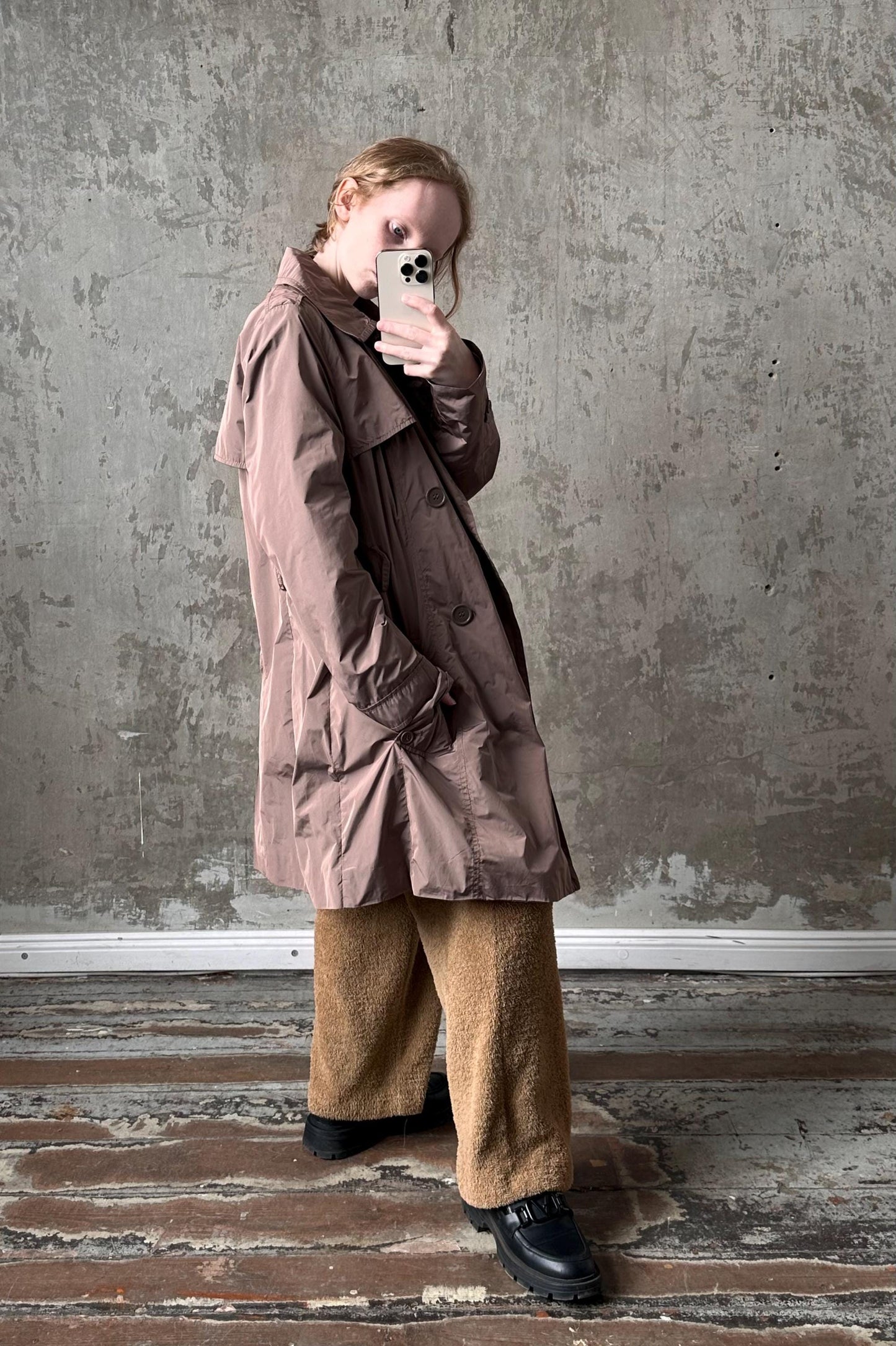 A person is taking a selfie with a smartphone, wearing a brown coat and beige pants, standing in front of a concrete wall.