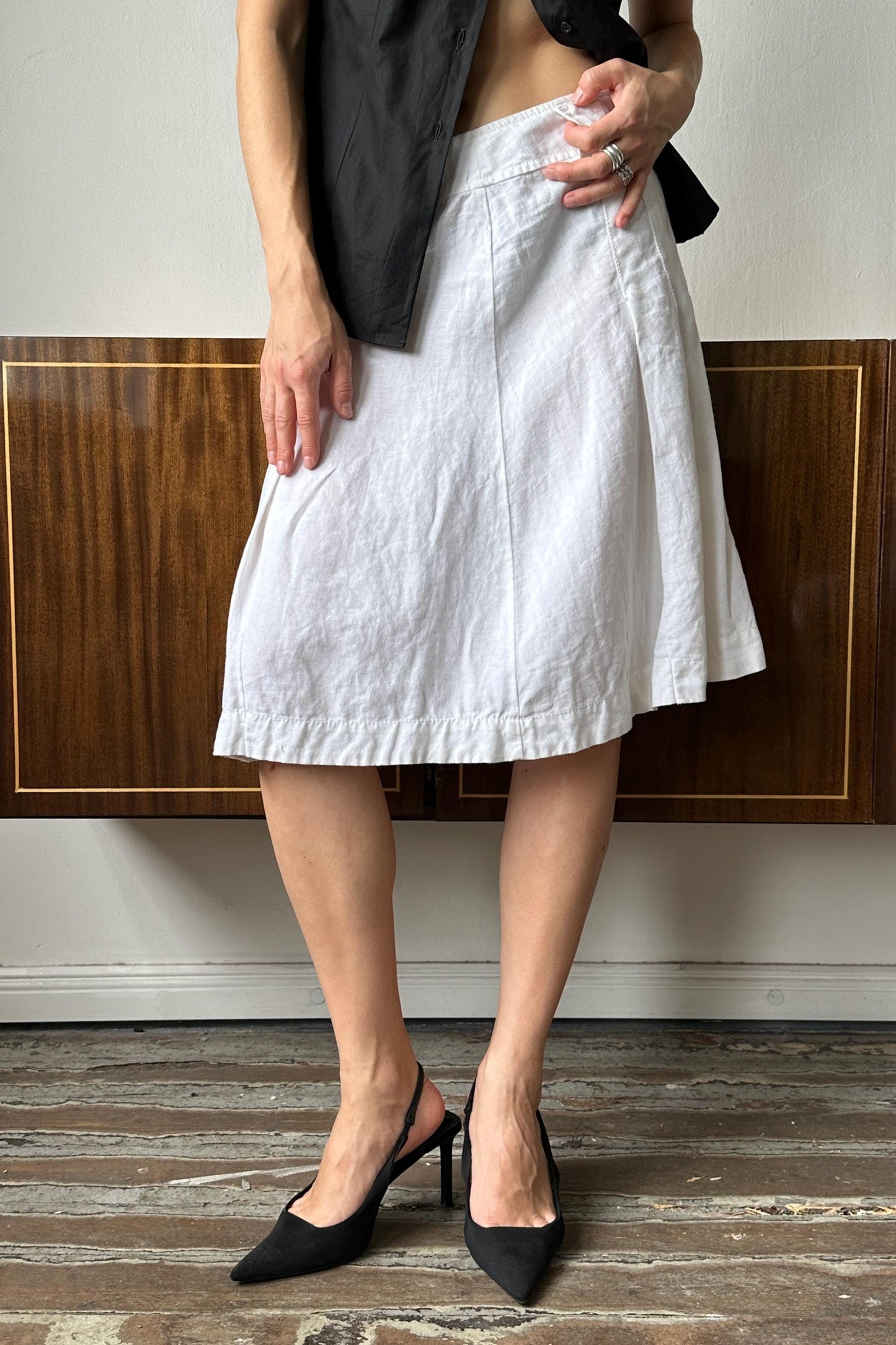 A person wearing a white skirt and black heels stands in front of a wooden cabinet.