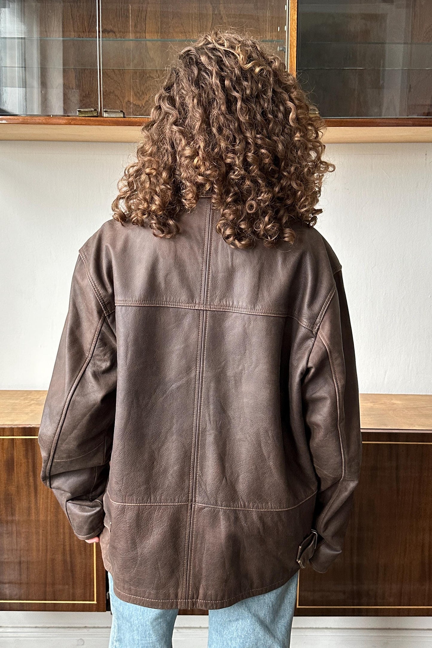 a person with curly hair wearing a brown leather jacket, standing in front of a glass display case.