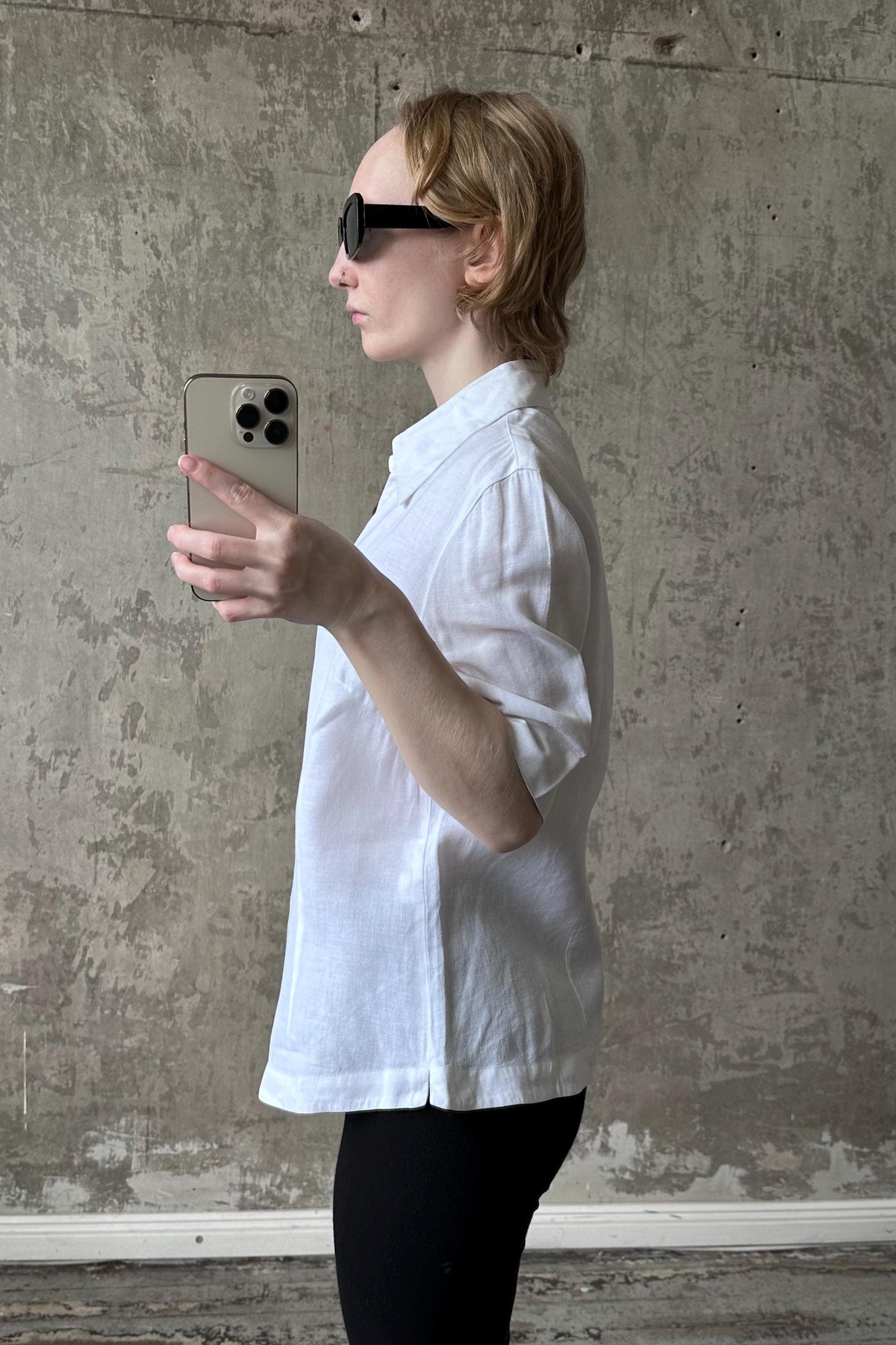 A person wearing a white shirt and sunglasses is holding a smartphone and taking a selfie in front of a concrete wall.