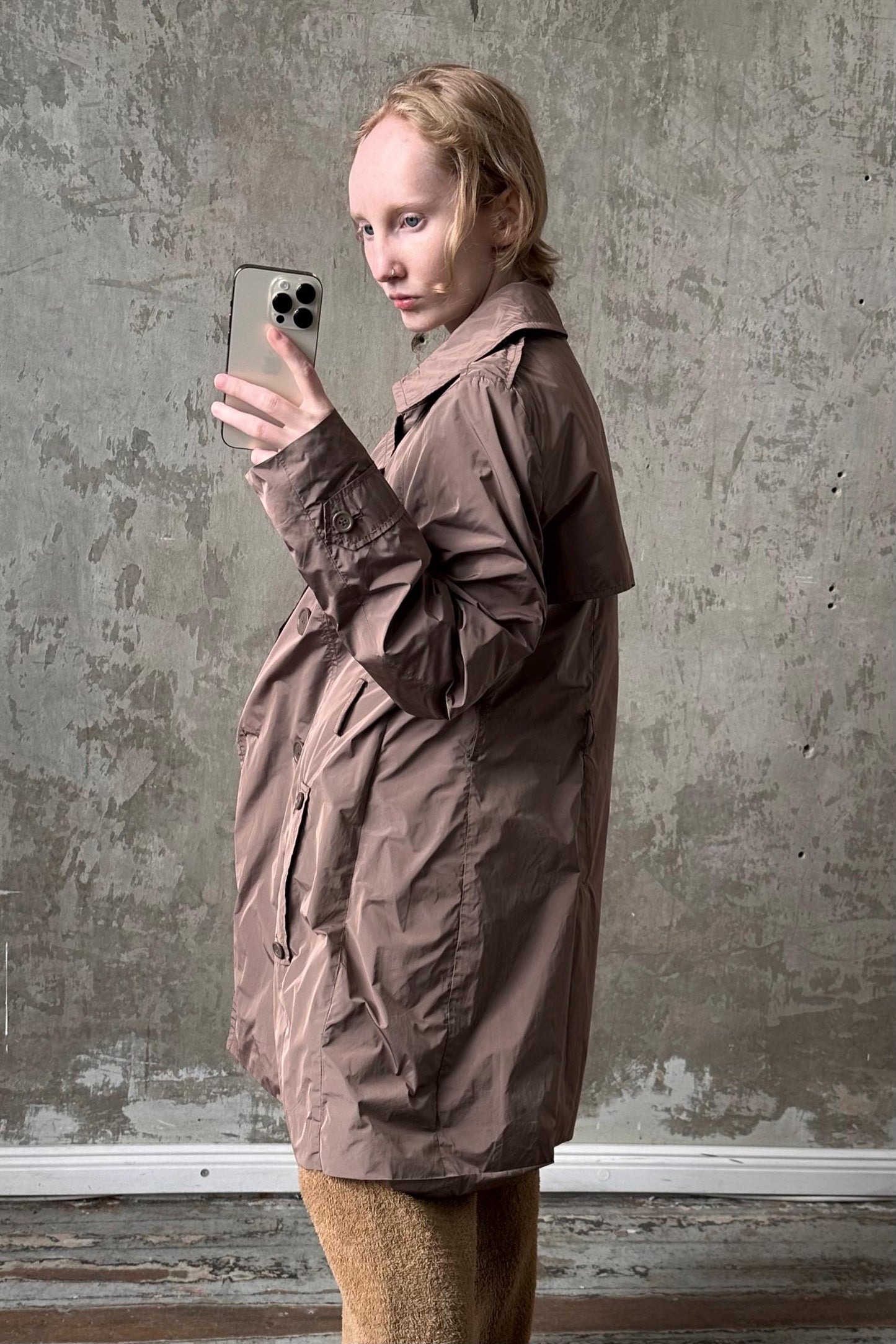 A person in a brown coat is holding a smartphone and taking a selfie in front of a concrete wall.