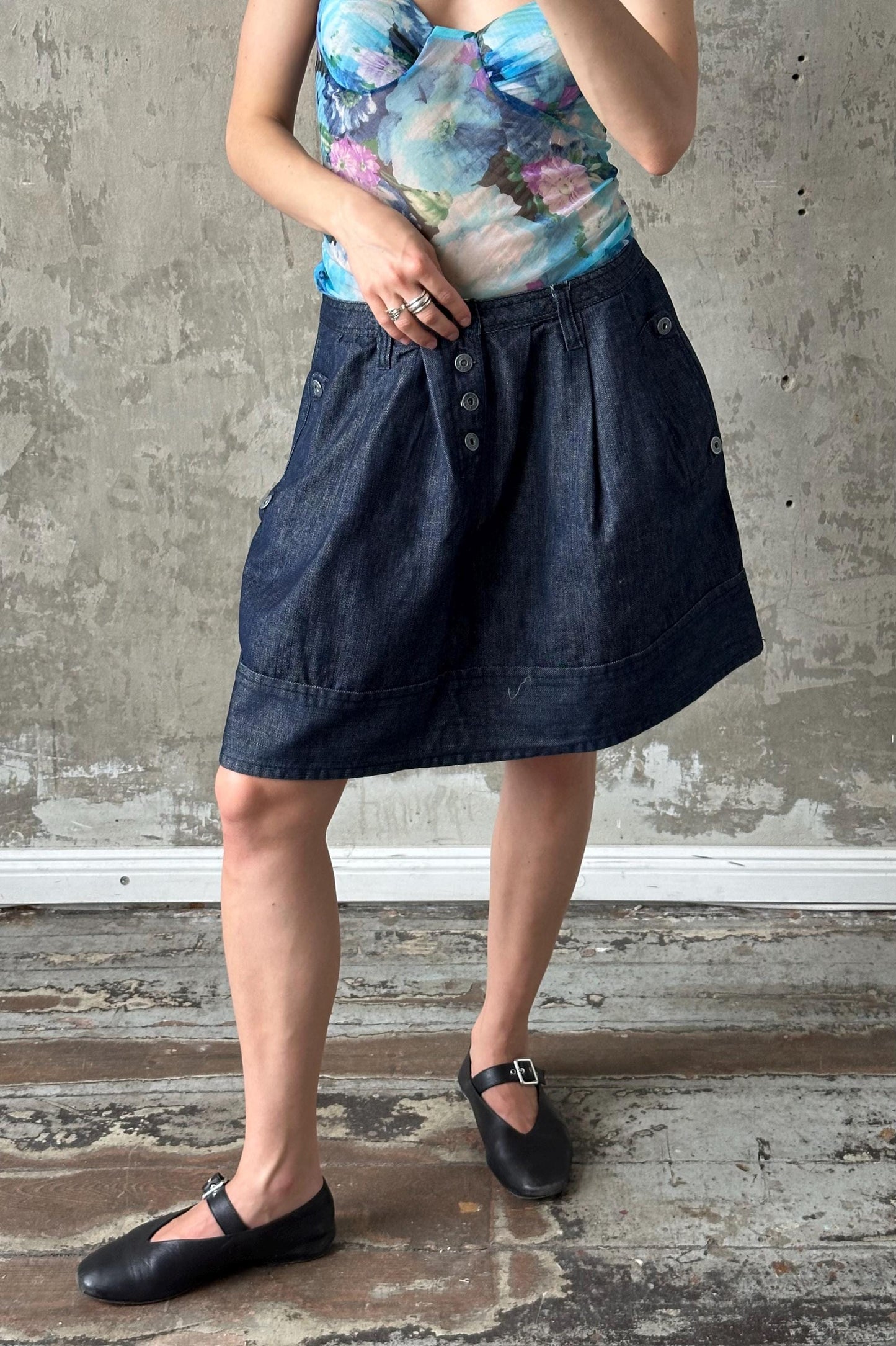 A woman wearing a floral top and a denim skirt stands in front of a concrete wall, with her hands in her pockets.