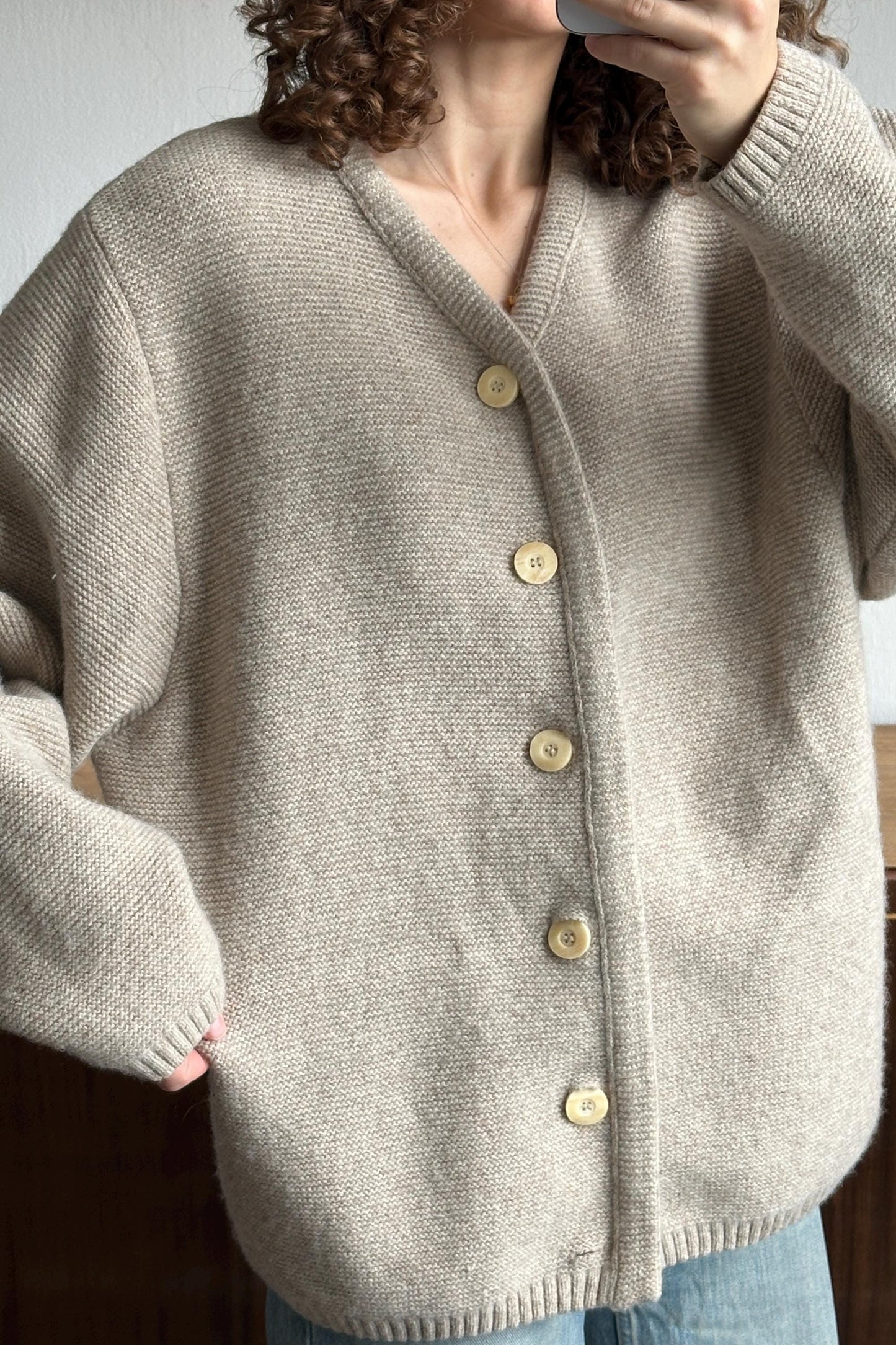 A person wearing a beige cardigan with gold buttons, taking a selfie with their phone.
