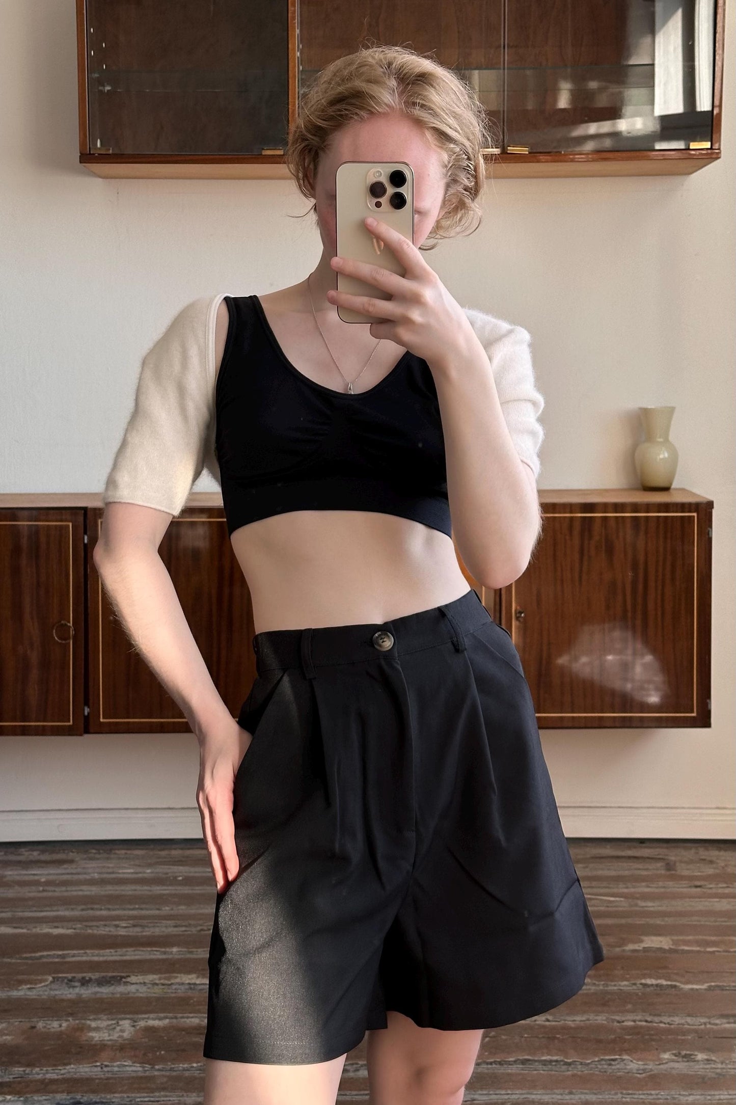 A woman is taking a selfie with her phone, wearing a black crop top and black shorts, and standing in front of a wooden cabinet.