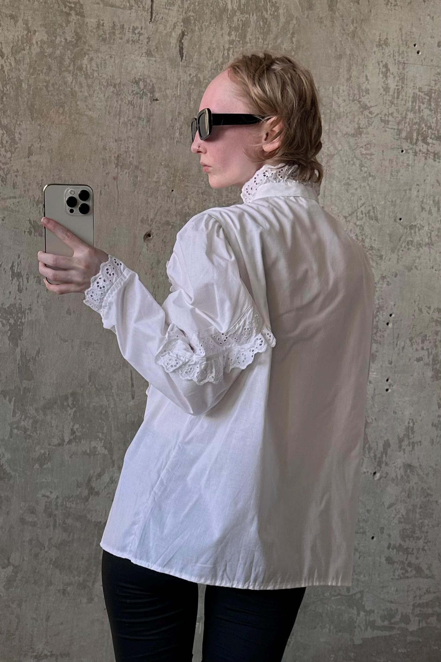 A woman wearing a white blouse and sunglasses is holding a smartphone and taking a selfie in front of a concrete wall.