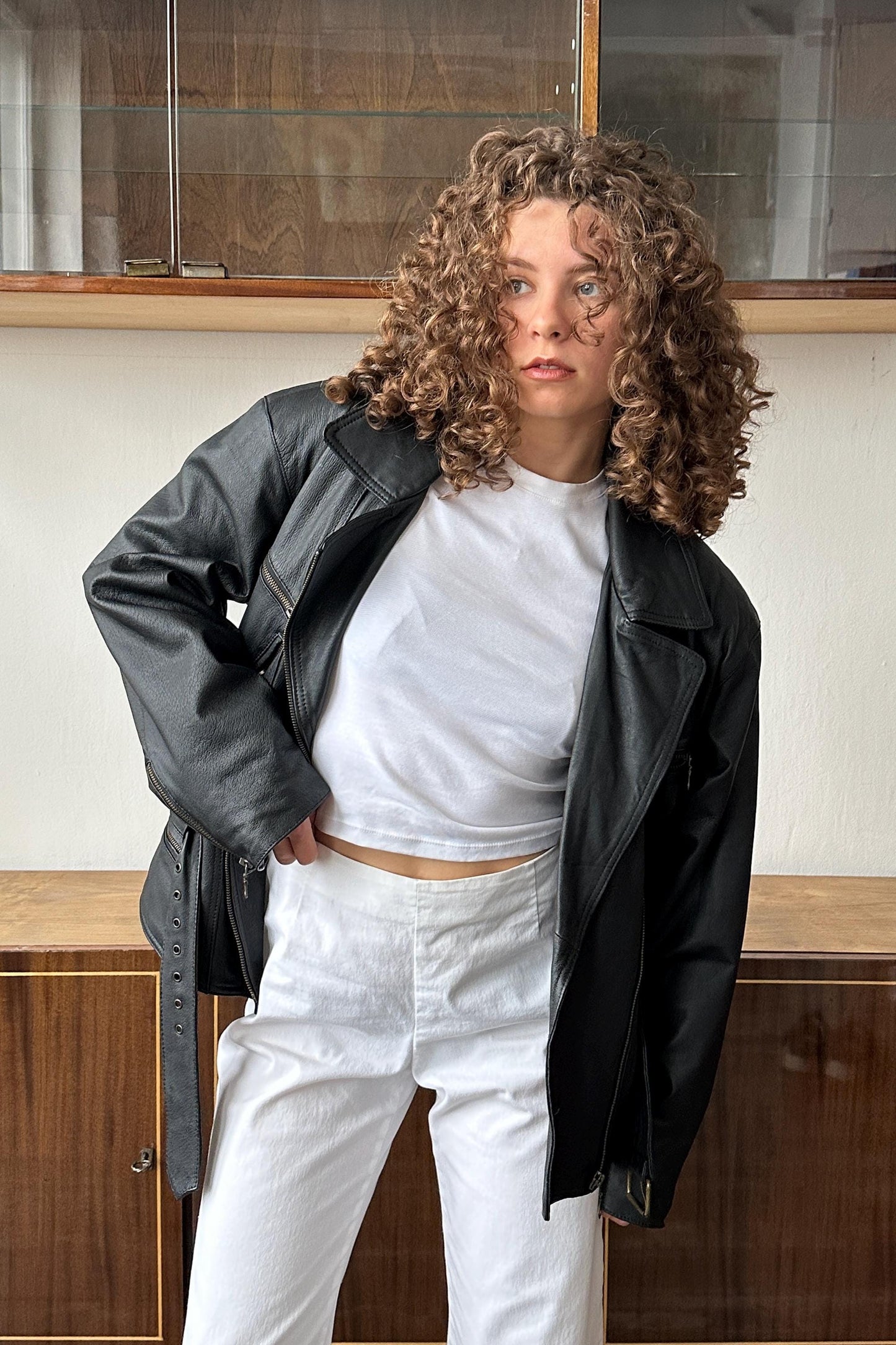 A woman with curly hair wearing a black leather jacket and white pants stands in front of a wooden cabinet.