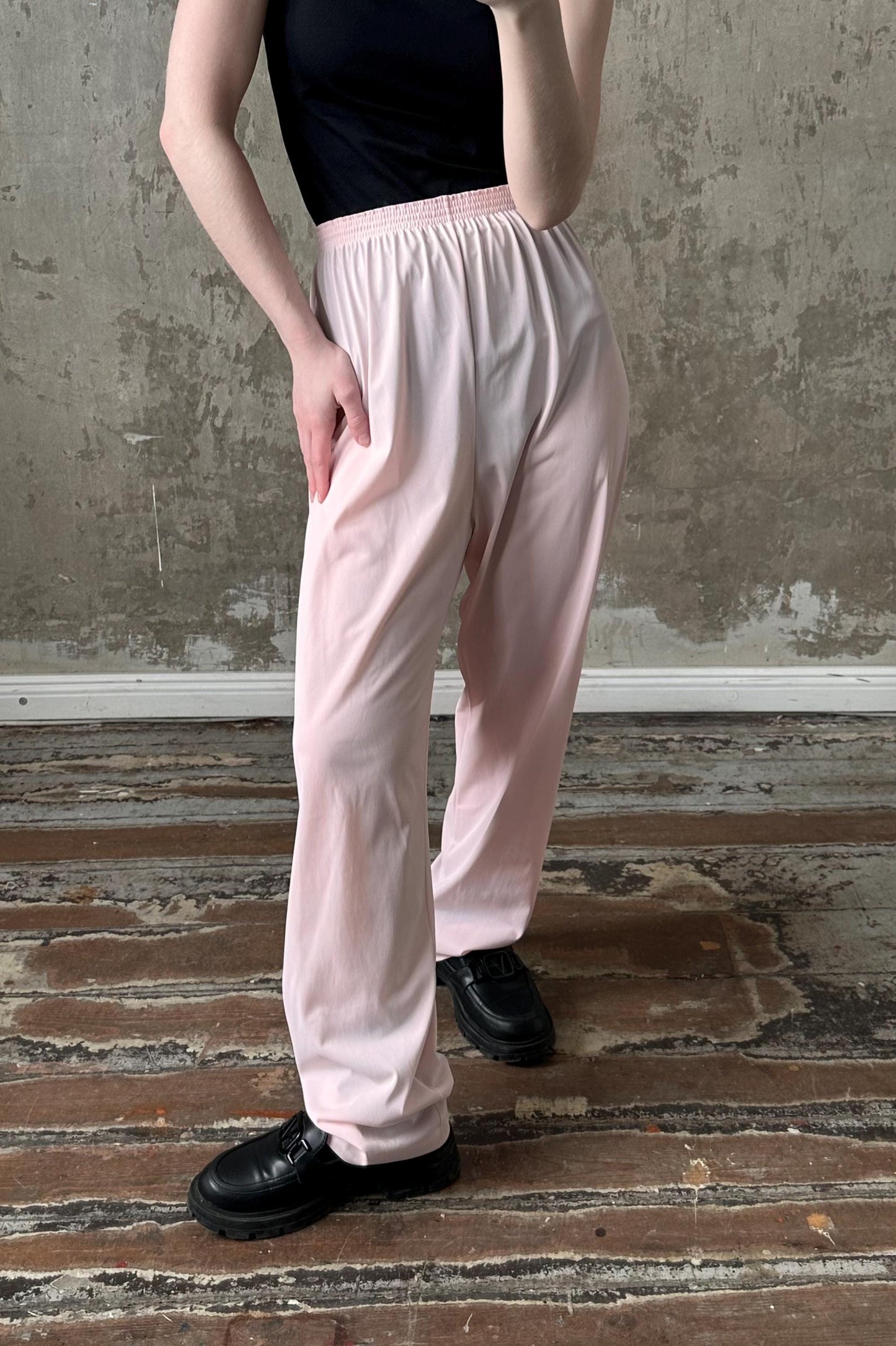 A person wearing a black top and pink pants is standing in front of a concrete wall.