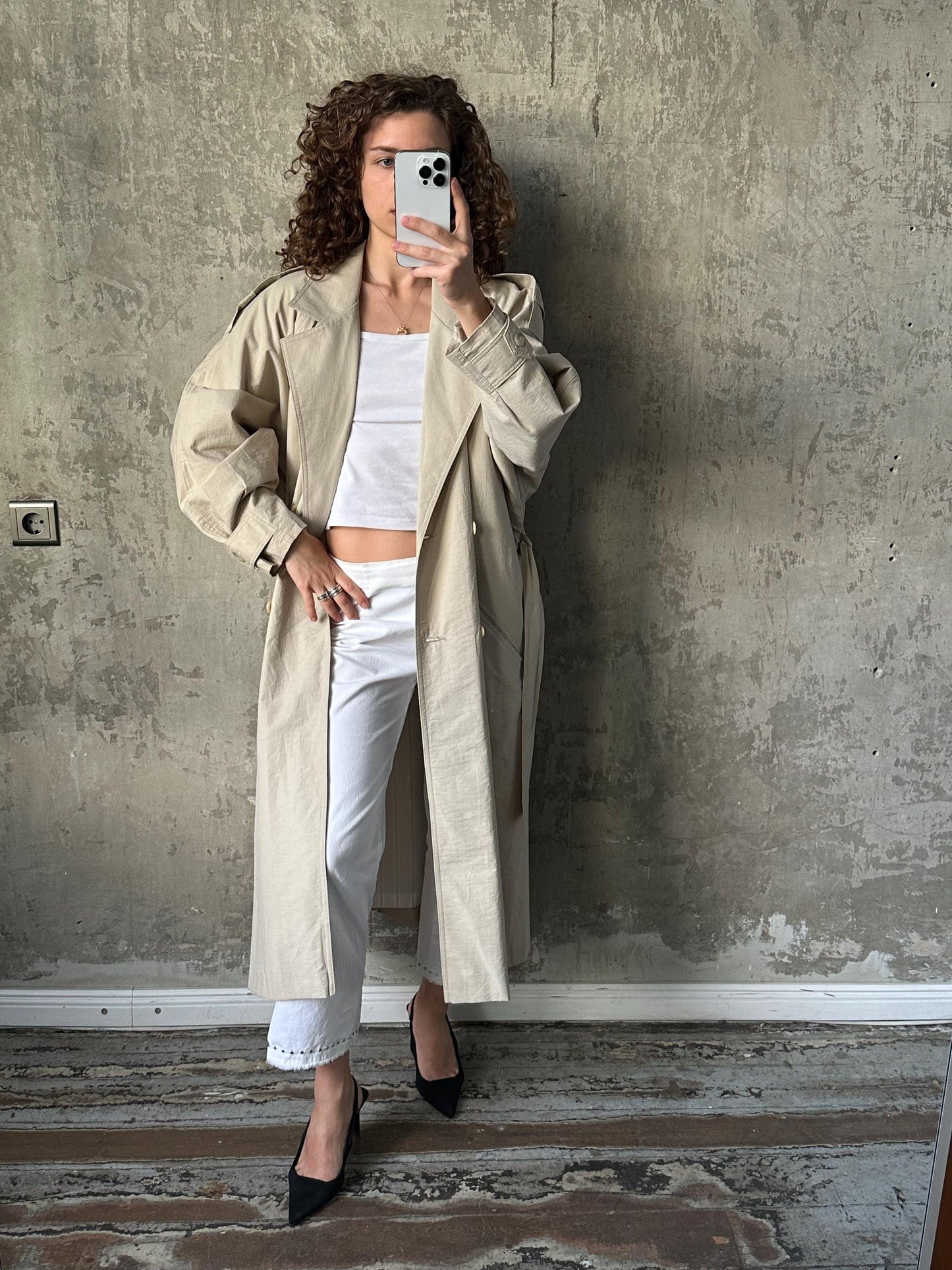 A woman in a beige trench coat and white pants is taking a selfie with a smartphone, standing against a concrete wall.