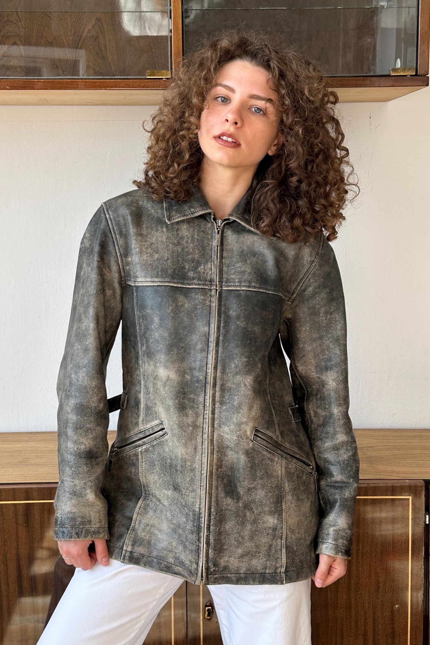 A woman with curly hair wearing a gray leather jacket stands in front of a wooden cabinet.