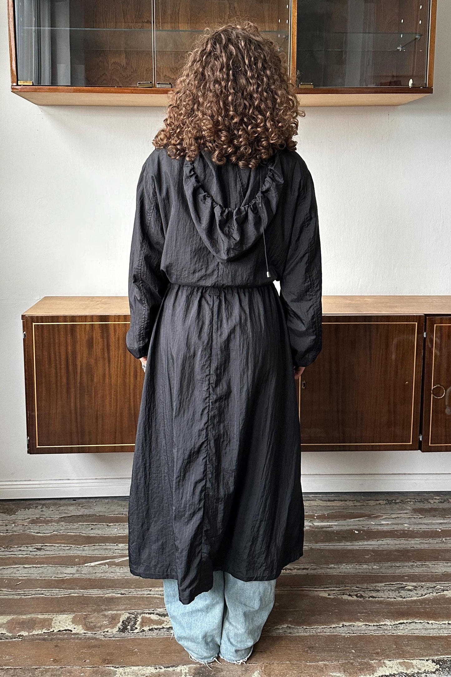 A person with curly hair wearing a long black coat stands in front of a wooden cabinet.