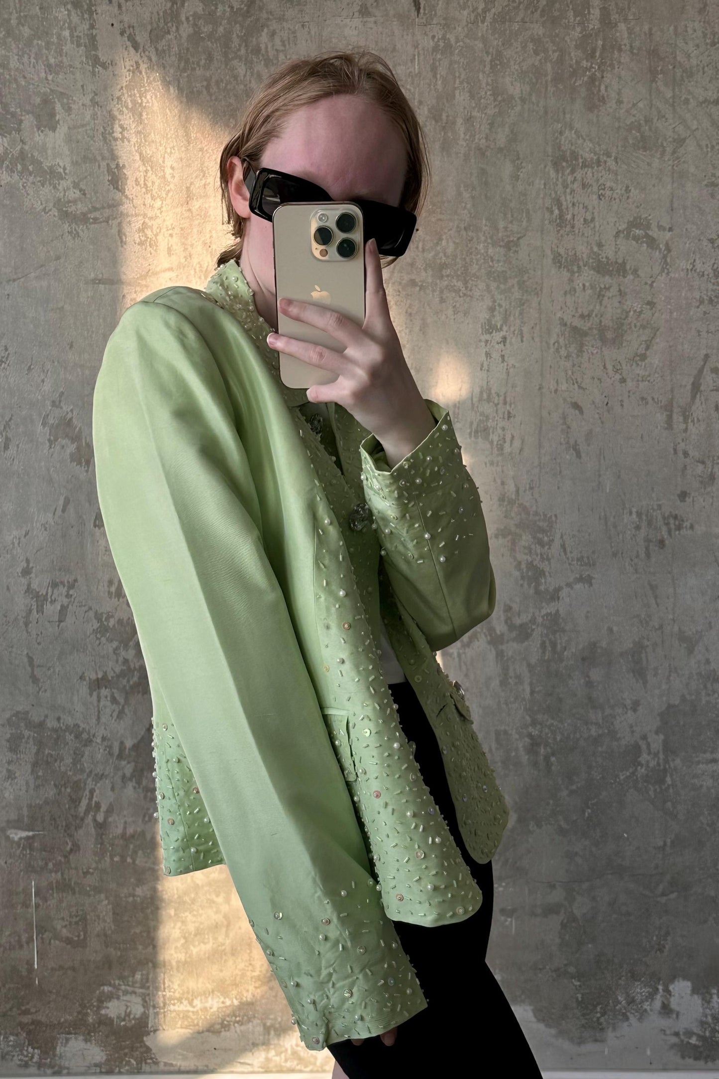A person is taking a selfie with a green jacket and sunglasses, holding a smartphone in front of their face.