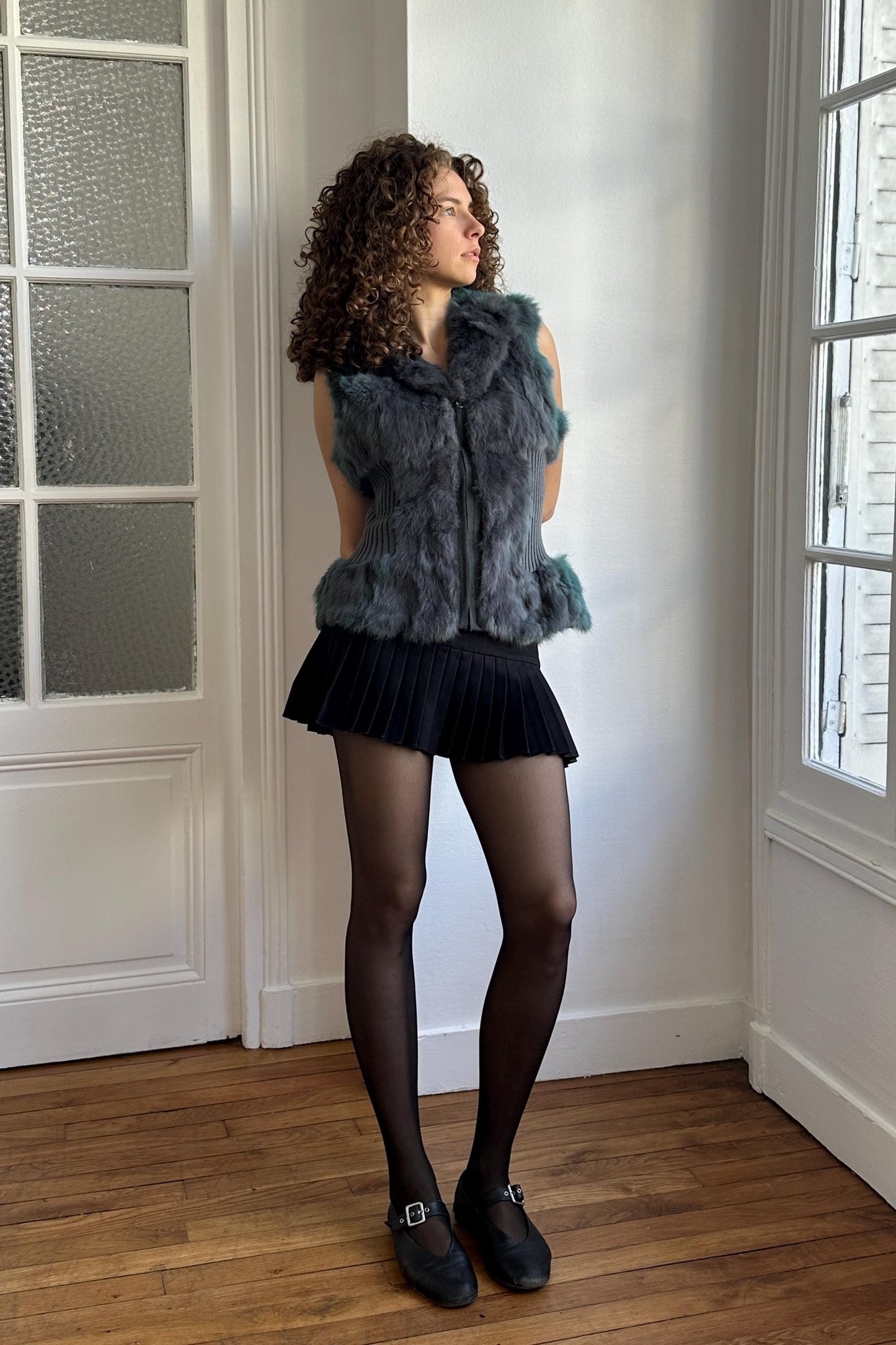 A woman with curly hair stands in a room, wearing a gray fur vest, black skirt, and black shoes.