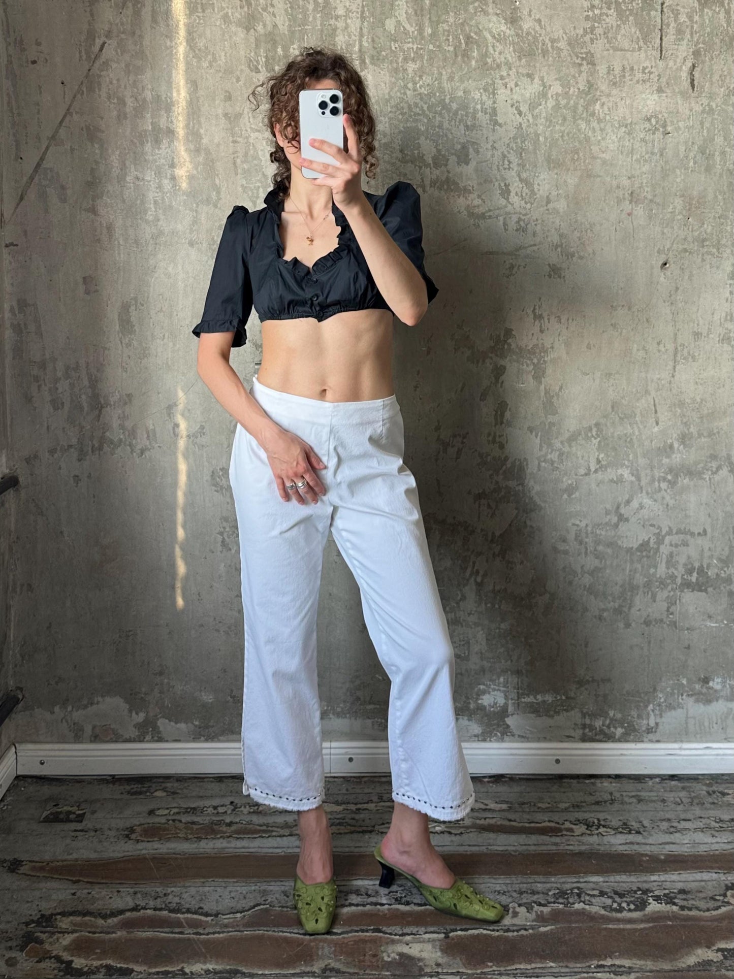 A woman is taking a selfie with her phone, standing in front of a concrete wall. She is wearing a black top, white pants, and green shoes.