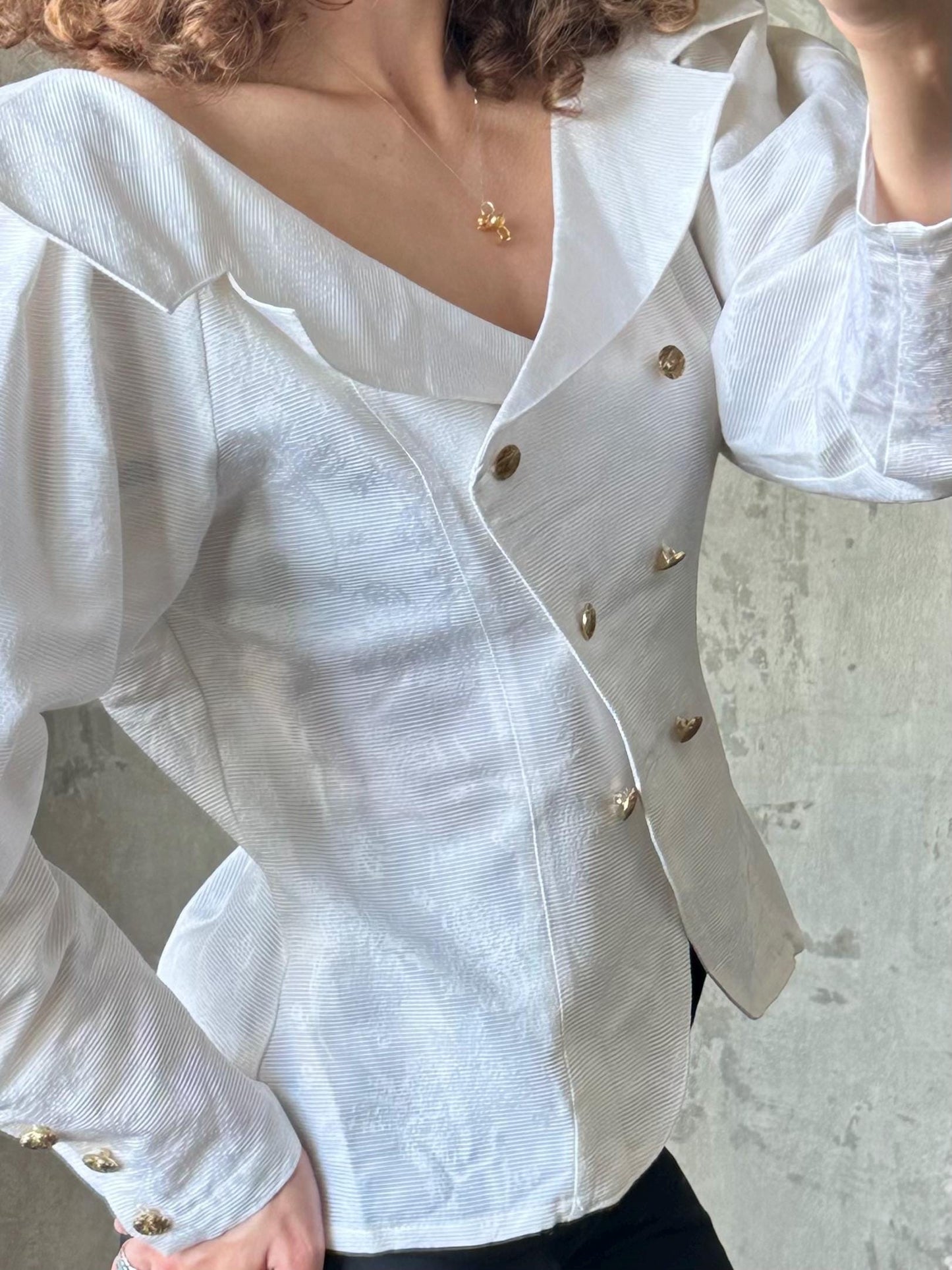 A close-up view of a person wearing a white blouse with a high collar and buttons, standing against a concrete wall.