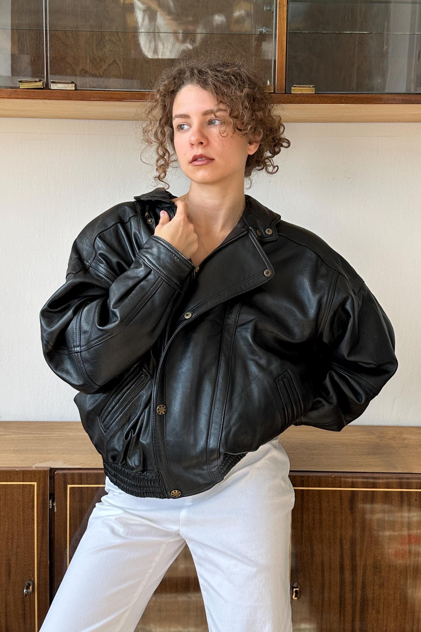 A woman wearing a black leather jacket stands in front of a wooden cabinet.