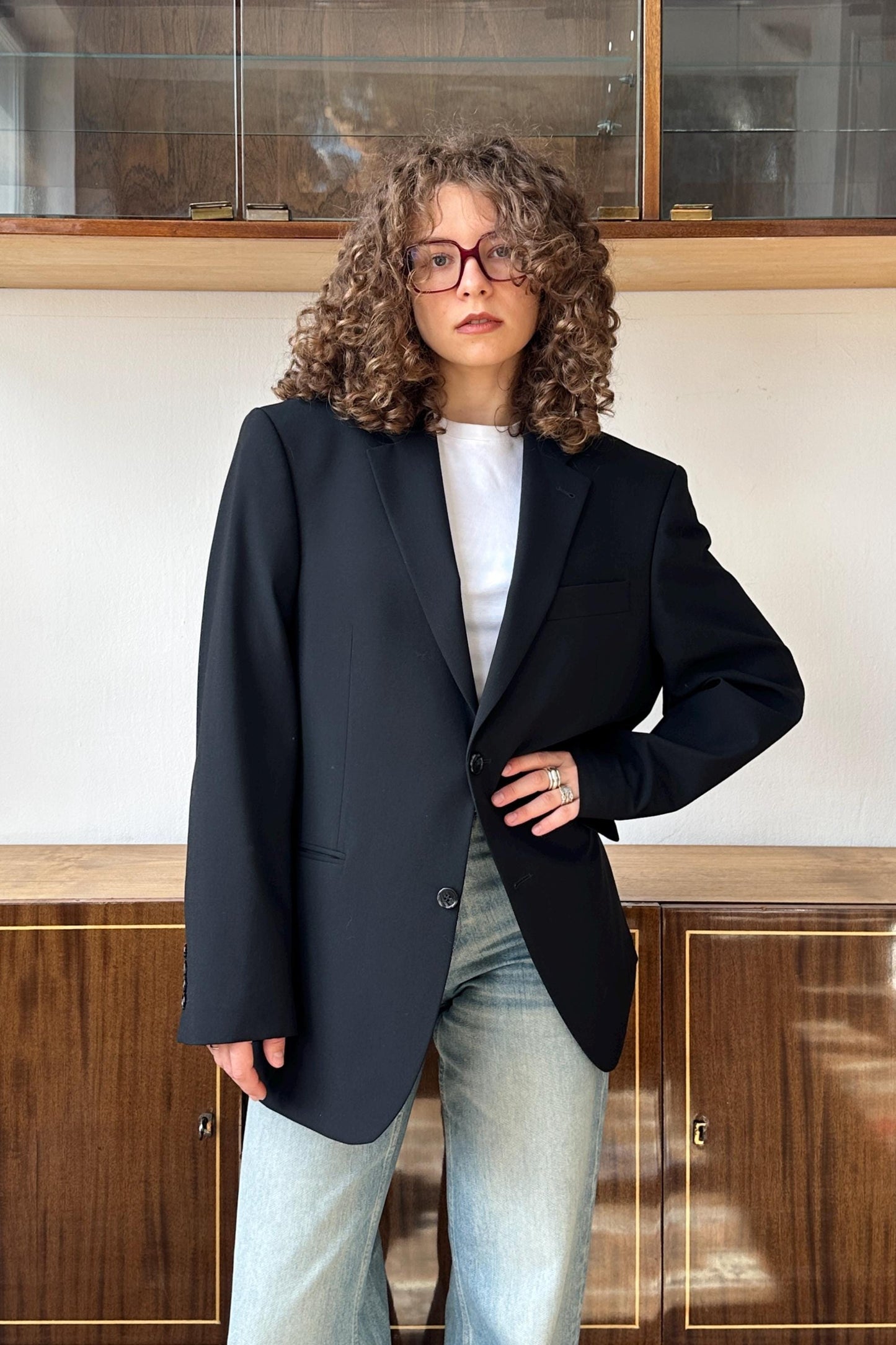 A woman with curly hair wearing a black blazer and jeans stands in front of a wooden cabinet.