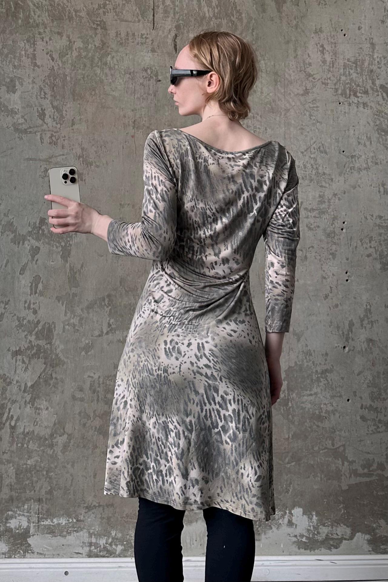 A woman wearing a leopard print dress is holding a smartphone and taking a selfie in front of a concrete wall.
