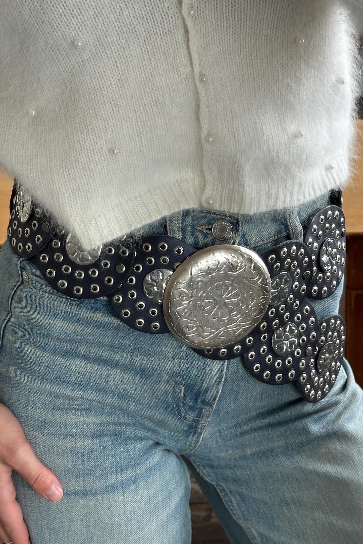A close-up of a person wearing a white sweater and blue jeans, with a large, decorative belt featuring a metal buckle.