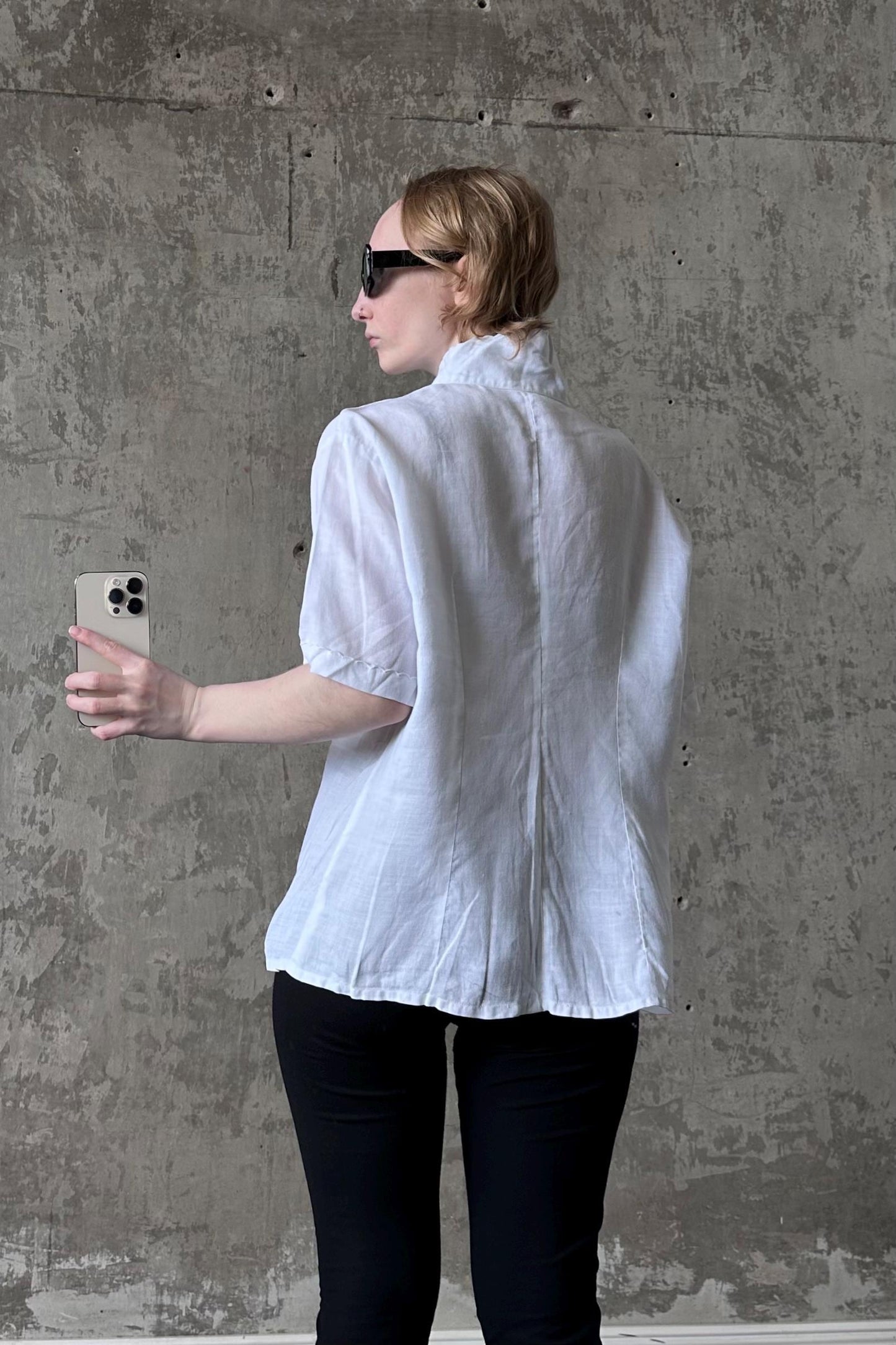 A woman in a white blouse and black pants is holding a smartphone and looking at it, standing in front of a concrete wall.