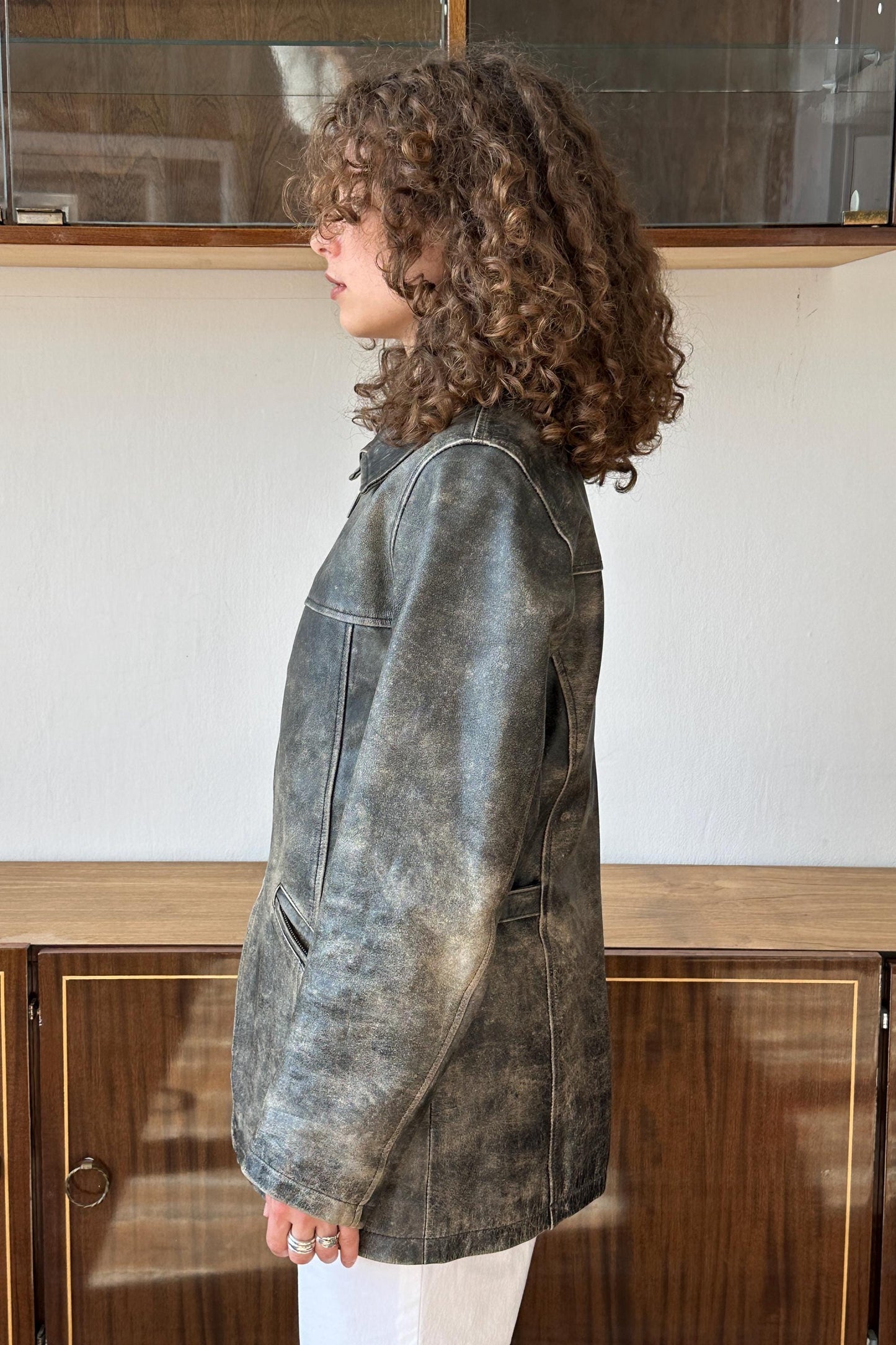 A woman with curly hair wearing a gray leather jacket stands in front of a wooden cabinet.