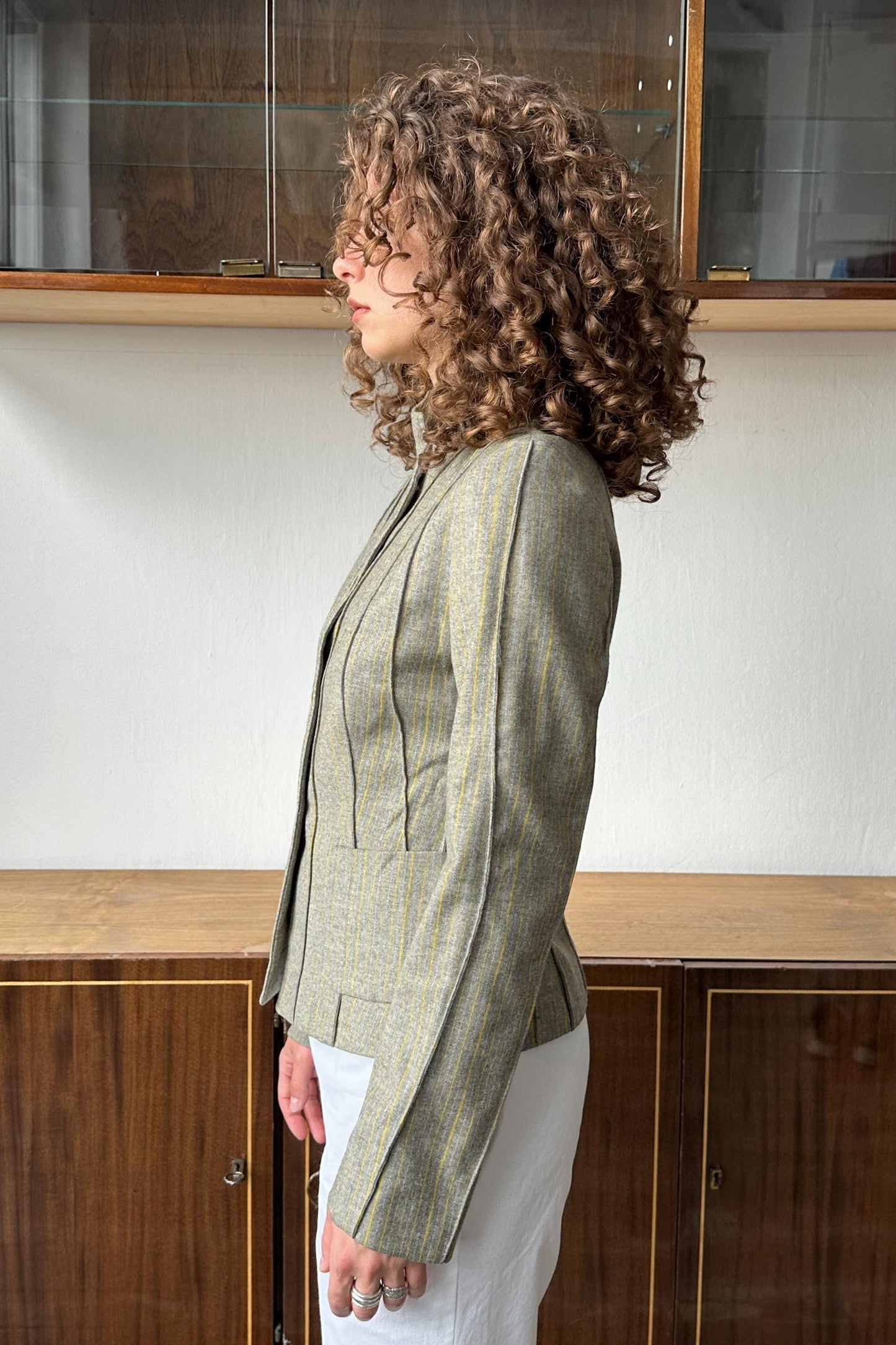A woman with curly hair wearing a striped jacket stands in front of a wooden cabinet.