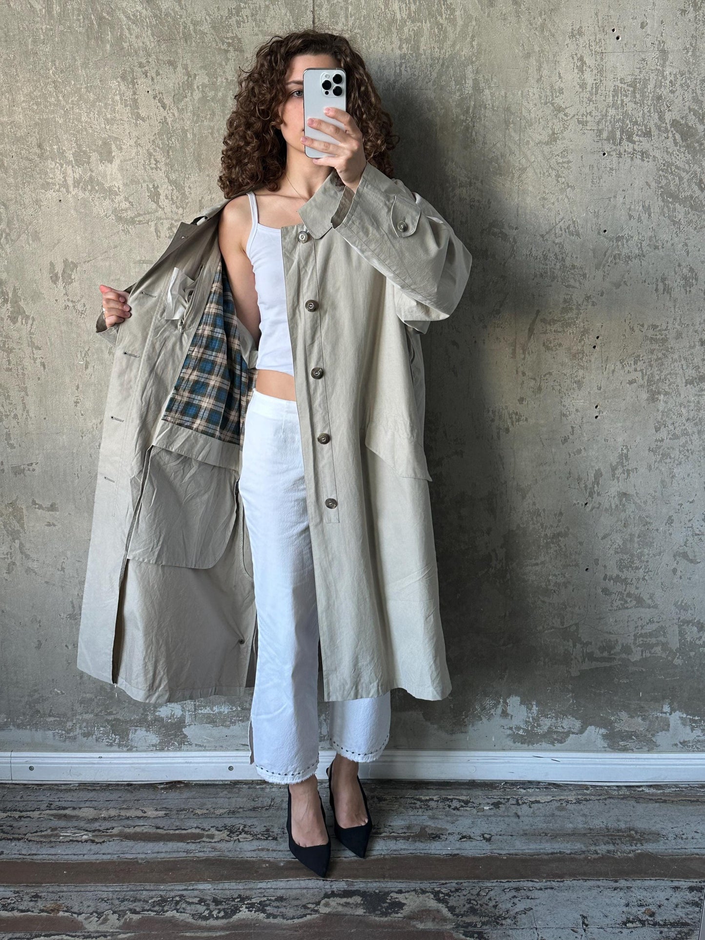A woman in a beige trench coat is taking a selfie with her phone against a concrete wall.