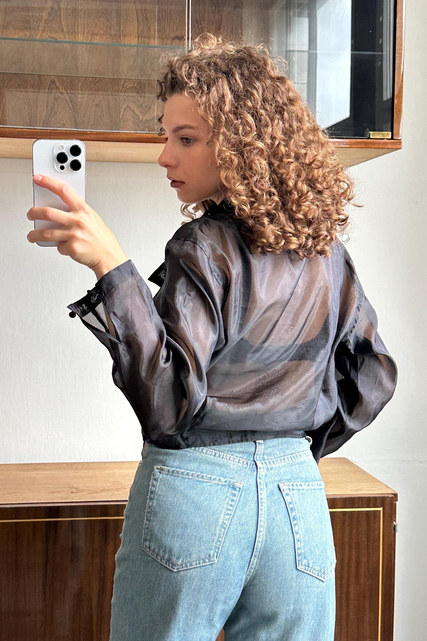 A woman with curly hair is taking a selfie with her phone, standing in front of a wooden cabinet.