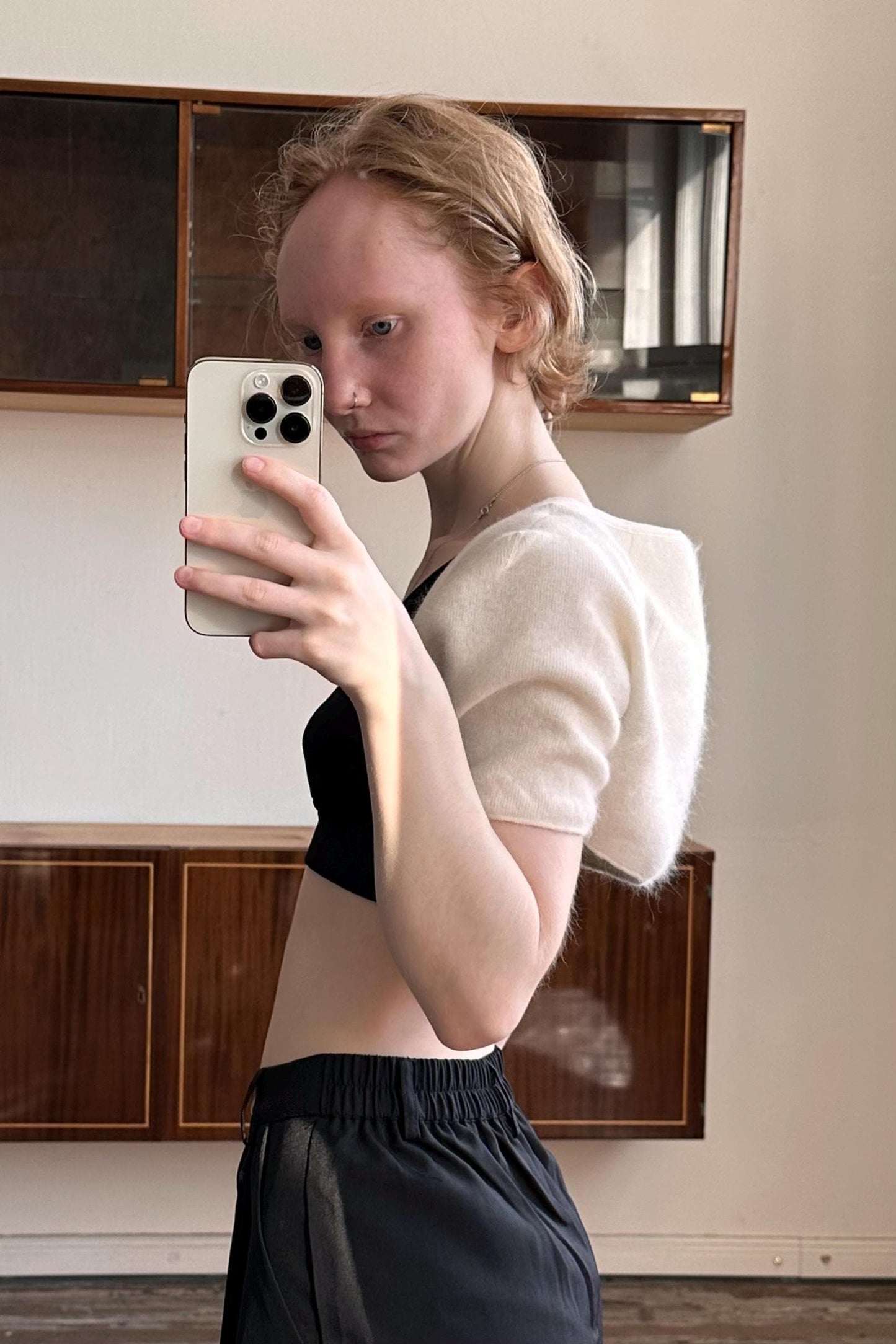 A young woman is taking a selfie with a white iPhone in a room with wooden cabinets.