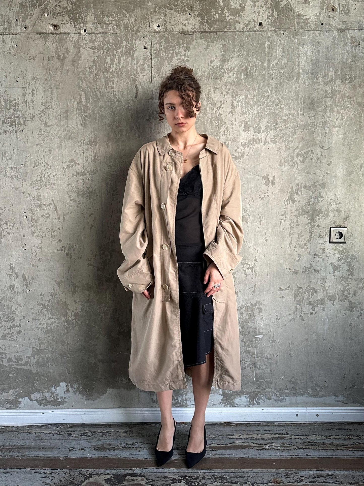 A woman wearing a beige trench coat and black dress stands against a concrete wall, looking directly at the camera.